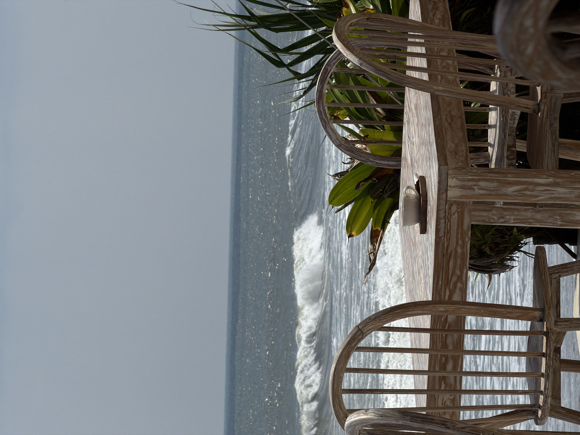 The image displays an outdoor dining or lounge area situated on a coastline in Belalang, Indonesia. A rectangular wooden table with a distressed light finish occupies the central foreground, surrounded by several matching wooden chairs featuring spindle backs. On the table, a white ceramic cup is placed on a small, dark wooden coaster, next to a small, dark rectangular object. Directly behind the table, a dense growth of green tropical foliage, appearing as large-leafed plants, provides a natural barrier. Beyond the foliage, the ocean extends to the horizon, characterized by active waves with visible white crests breaking across the dark blue-grey water. The sky above the ocean is a clear, light blue-grey, indicative of an overcast or hazy condition. No individuals are present in the scene.