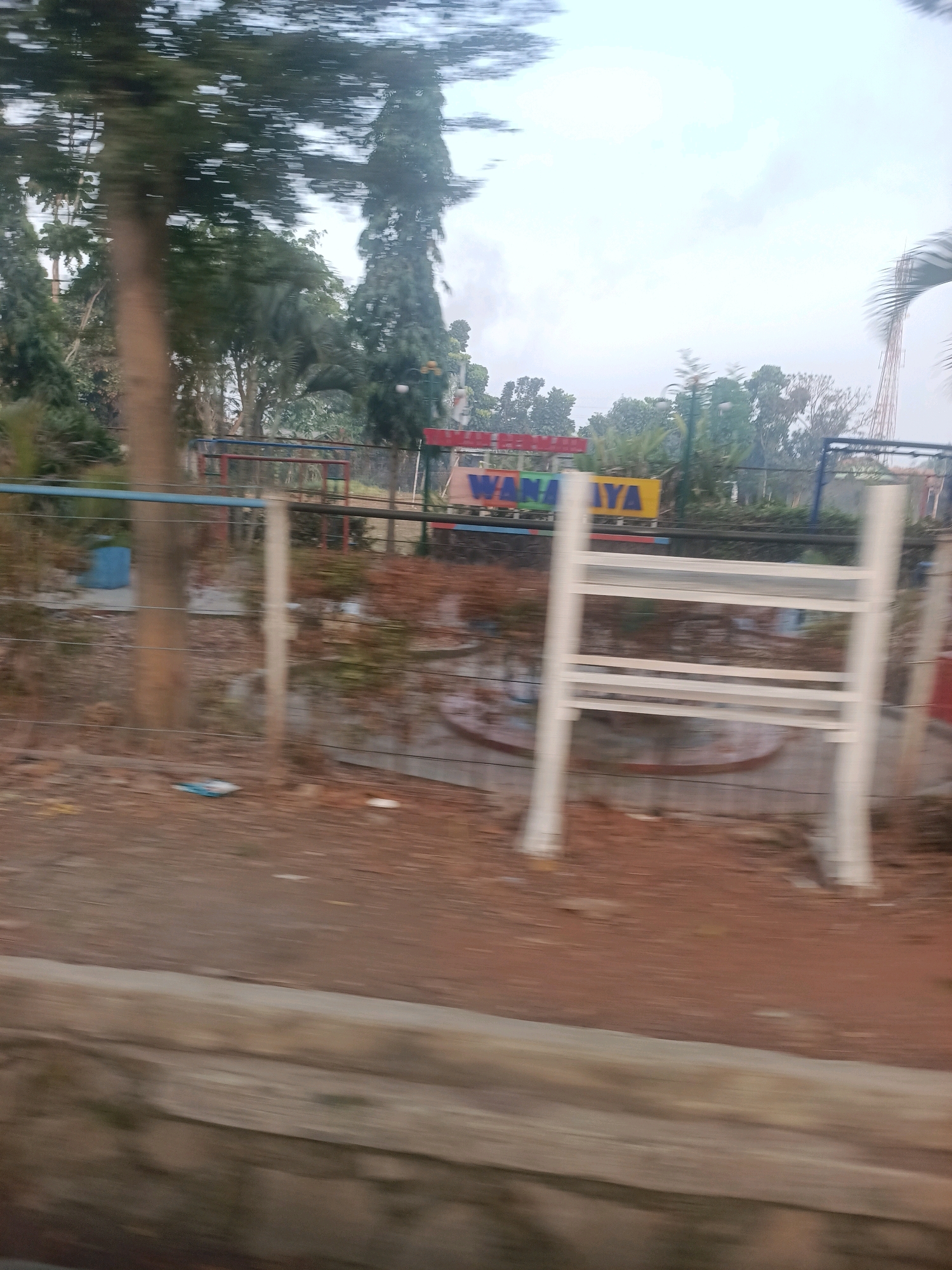 The image depicts a motion-blurred outdoor scene, likely a park or recreational area, with a portion of a city name visible. The name "WANAJAYA" is prominently displayed in colorful block letters on a yellow sign, suggesting the location is in Wanajaya, Indonesia. In the foreground, there is a light-colored metal structure, possibly playground equipment or seating, with a white railing. A wooden pole stands to the right of this structure. Behind it, a fence with horizontal blue and dark bars separates the immediate area from a dirt and dry foliage-covered ground. Trees of various sizes and types are visible in the background, some appearing tall and slender, others more bushy. A red-framed structure, possibly another piece of playground equipment, is partially visible behind the "WANAJAYA" sign. The sky is overcast and bright. The overall impression is of a public space on a cloudy day.