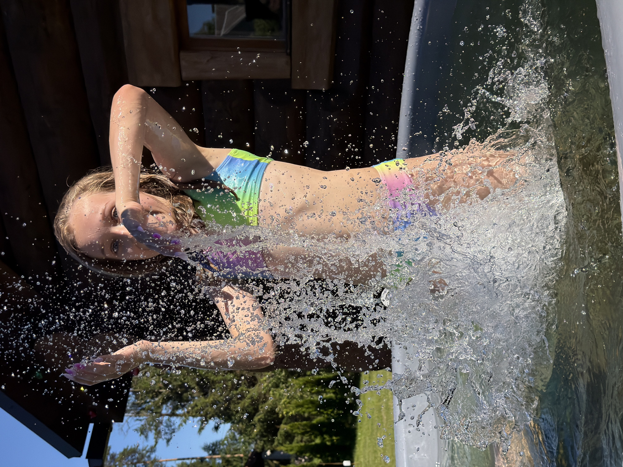 A young girl with blonde hair is captured mid-air, splashing into a body of water, likely a pool or hot tub. She is wearing a colorful bikini with a tie-dye pattern featuring shades of green, blue, pink, and purple. Her face shows a mixture of exhilaration and surprise, with her eyes wide open and her mouth slightly agape. Water droplets fly around her, frozen in motion by the camera's shutter speed, creating a dynamic and energetic effect.
The background suggests an outdoor setting on a sunny day. To the left, a rustic wooden structure, possibly a cabin or sauna, is visible. Trees and a bright blue sky can be seen in the distance, indicating it's likely daytime and the weather is fair. The overall impression is one of carefree summer fun and a moment of pure joy. No visible text is present in the image.