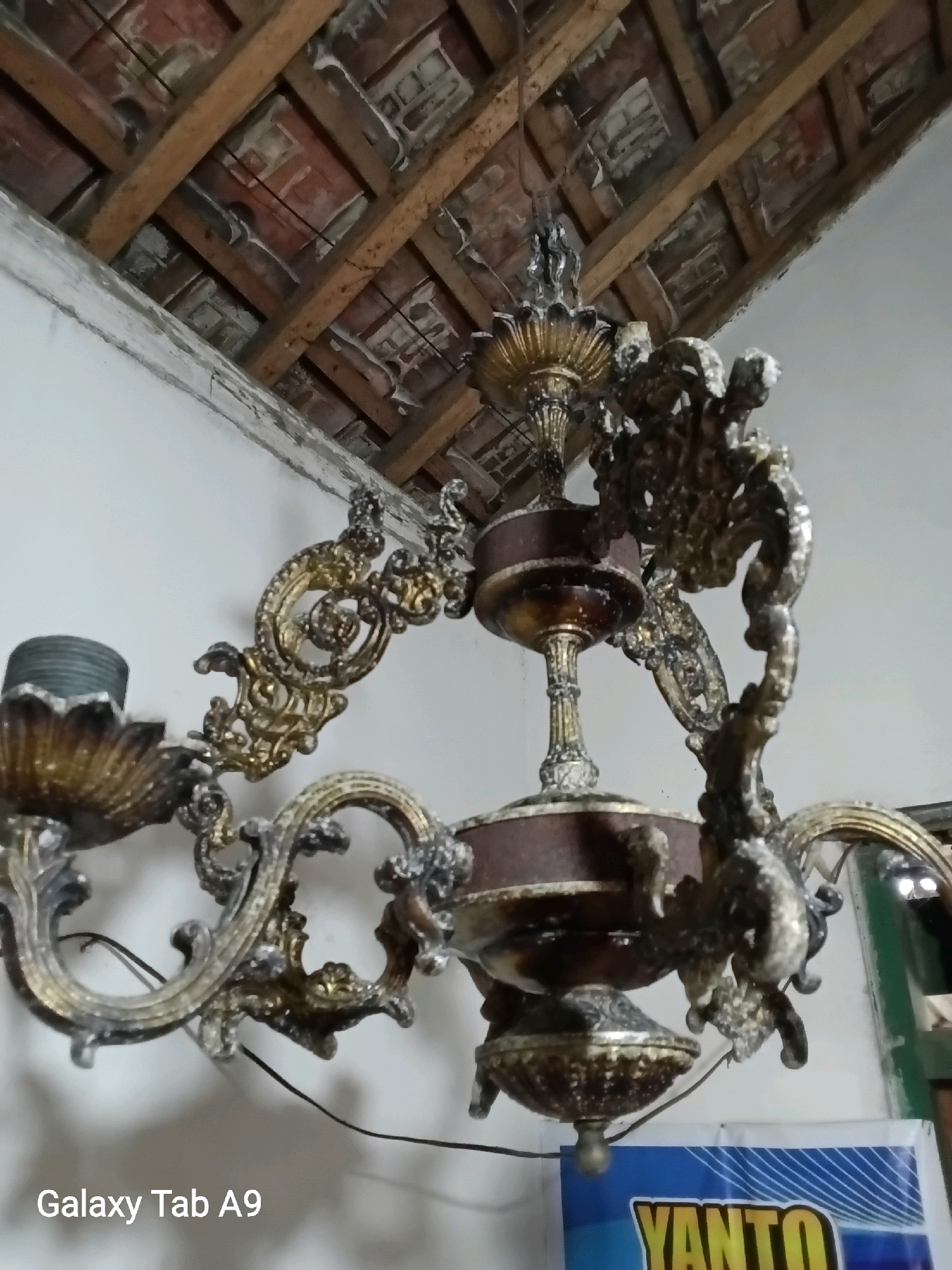 The image depicts an ornate, antique chandelier hanging from a wooden beam ceiling. The chandelier is made of a dark metal, likely brass or bronze, with intricate scrolling details and decorative elements. It appears to have multiple arms, each designed to hold a light bulb. The ceiling is constructed with exposed wooden beams and red tiles, suggesting an older building. The walls are painted white. In the foreground, a portion of a blue and yellow banner with the text "YANTO" is visible. The watermark "Galaxy Tab A9" is in the bottom left corner. The scene is indoors, likely in a residential or historical setting within Karangan, Indonesia.
