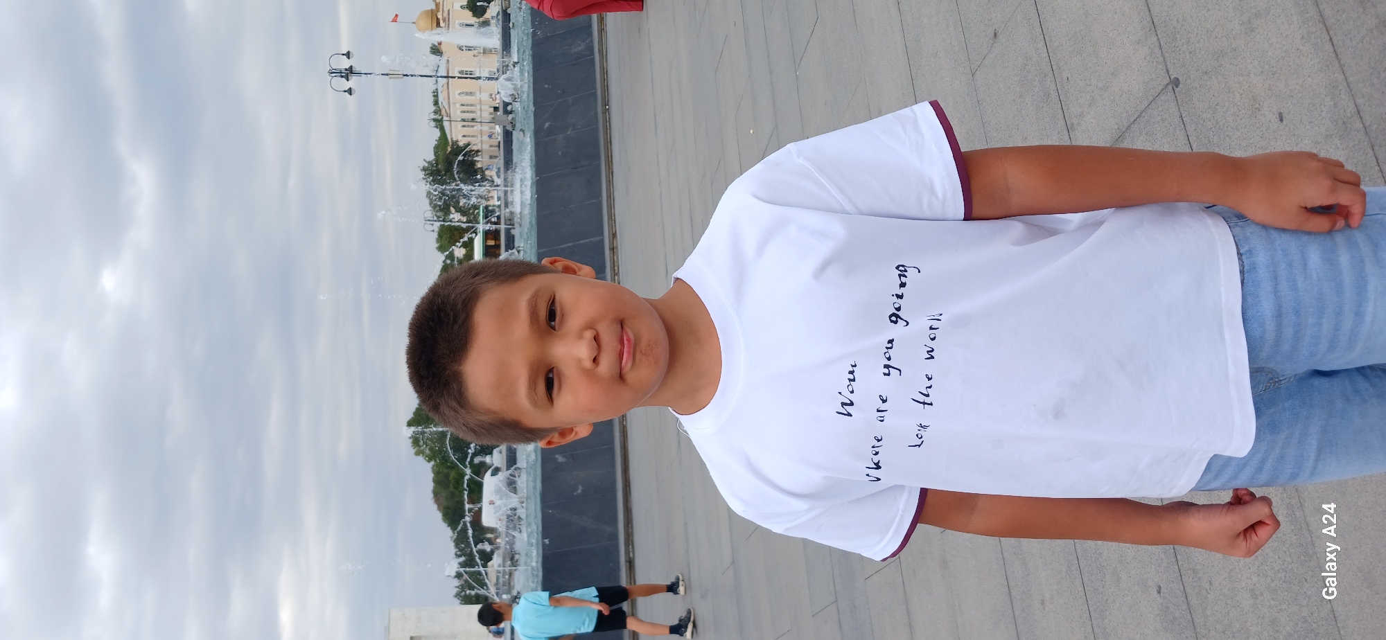 A young boy stands in the foreground, looking directly at the camera with a neutral expression. He is wearing a white t-shirt with a dark, handwritten message that appears to read "Wow, Where are you going? Let the world." He is also wearing light blue denim shorts. In the background, a fountain is visible with water spraying upwards. Behind the fountain, there are buildings and trees, suggesting an urban park or public square. Another young boy in a blue t-shirt and black shorts is walking away from the camera in the middle ground, to the left of the main subject. The sky is overcast with clouds. The location is the Ala-Too Square in Bishkek City, Kyrgyzstan, as indicated by the prominent fountain and architecture. The paving stones of the square are visible in the foreground. The overall scene depicts a casual outdoor setting on an overcast day.