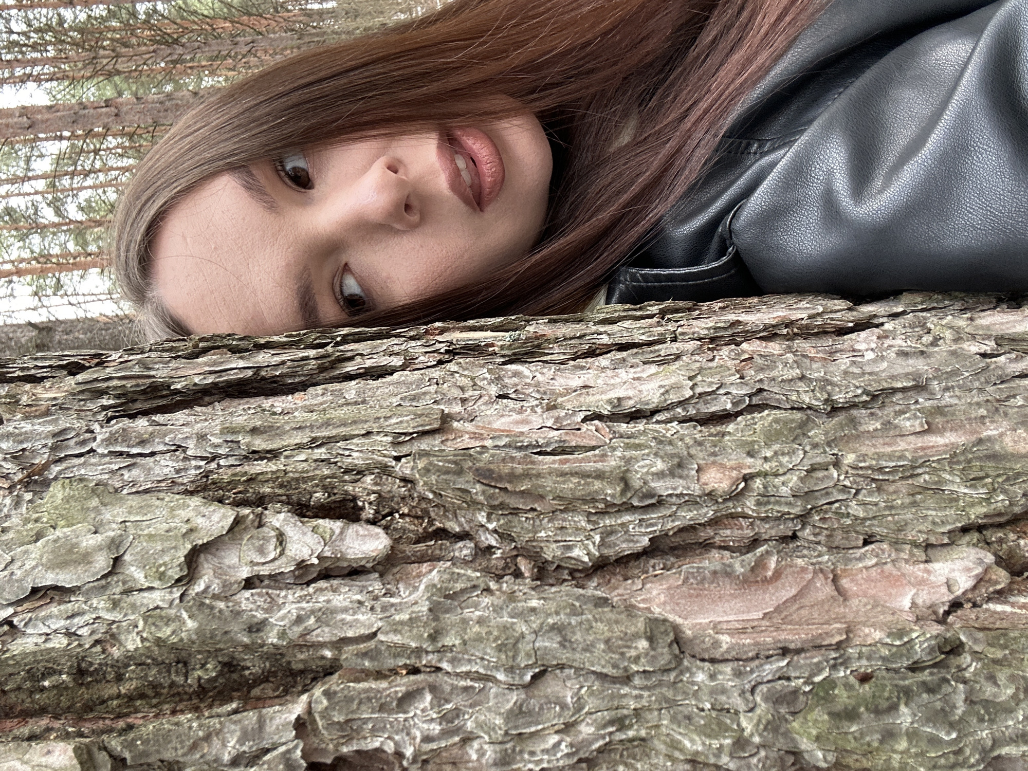 A young woman with long, brown hair and wearing a black leather jacket is lying on a tree trunk in a wooded area. Her head is tilted, and she is looking up at the camera. The bark of the tree is rough and textured. Thin trees and sky appear in the background. The location is in a wooded area, possibly near Chaikovsky, Russia.