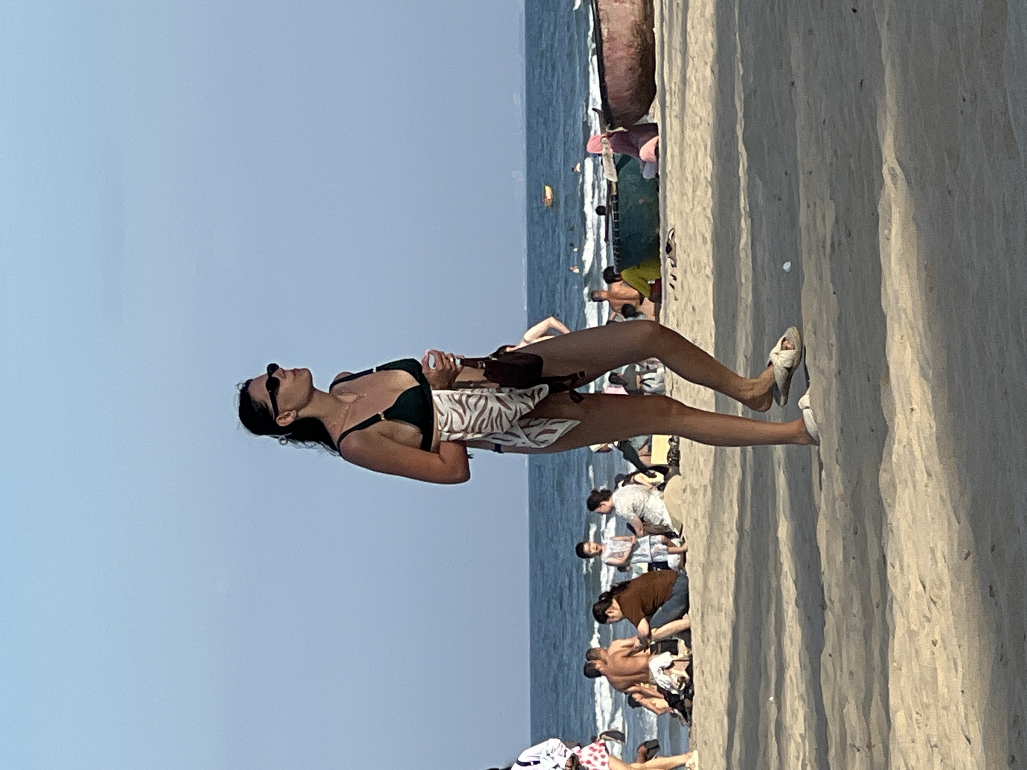 The image captures a beach scene, likely in Da Nang, Vietnam, with the ocean in the background and a sandy shoreline in the foreground. A woman in a dark green bikini and sunglasses is walking towards the right side of the frame, holding a patterned towel over her shoulder and a small bag. She is wearing white sandals. Behind her, a crowd of people are visible on the beach, some sitting and others standing, with the ocean waves gently rolling in. Several boats, including a traditional round boat and a larger wooden vessel, are present on the sand and in the water. The sky is clear and blue.