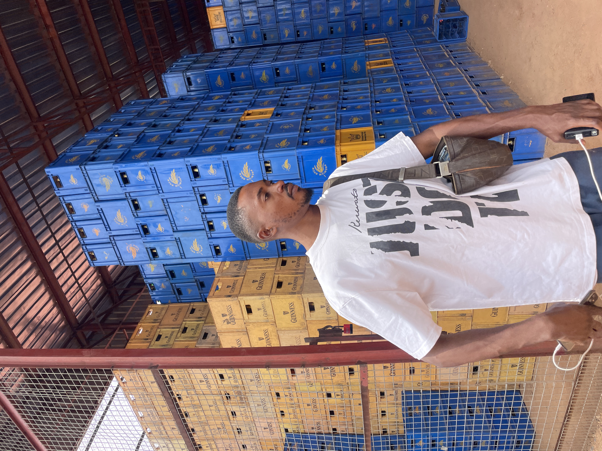 A man stands inside a warehouse or storage facility in Gura Top, Nigeria. He is wearing a white t-shirt with black lettering and a brown messenger bag, holding a phone. He is positioned in front of stacks of plastic crates filled with Guinness beer bottles. Most of the crates are blue with yellow labels. Some of the crates behind him are yellow with black labels. A wire fence runs along the bottom of the frame in front of the crates and the man. The ceiling is metal with brown support beams. The ground outside the fenced area appears to be dirt.