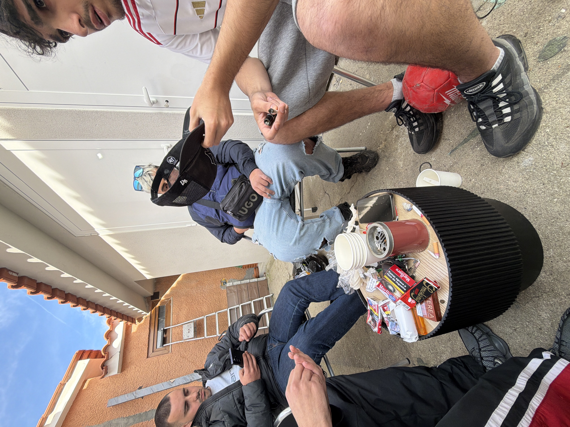 The image shows a group of young men gathered outdoors, likely in Vila Real, Portugal, as evidenced by Portuguese-language cigarette packaging visible on the table. The setting appears to be a backyard or patio. One man wears a white shirt with red stripes and a gold crest, and is seen holding a lighter. Another man, seated, wears a blue jacket over light blue jeans with a rip in the knee, along with a black cap and sunglasses. A third man is seen in a black jacket and dark jeans, holding what appears to be a mobile phone. A fourth man sits in gray sweatpants, with his foot resting on a red ball. A black table with a fluted edge is cluttered with cups, cigarette packs (including Marlboro), and other debris. In the background, part of a building with an orange tile roof and a ladder is visible. The scene suggests a casual social gathering.