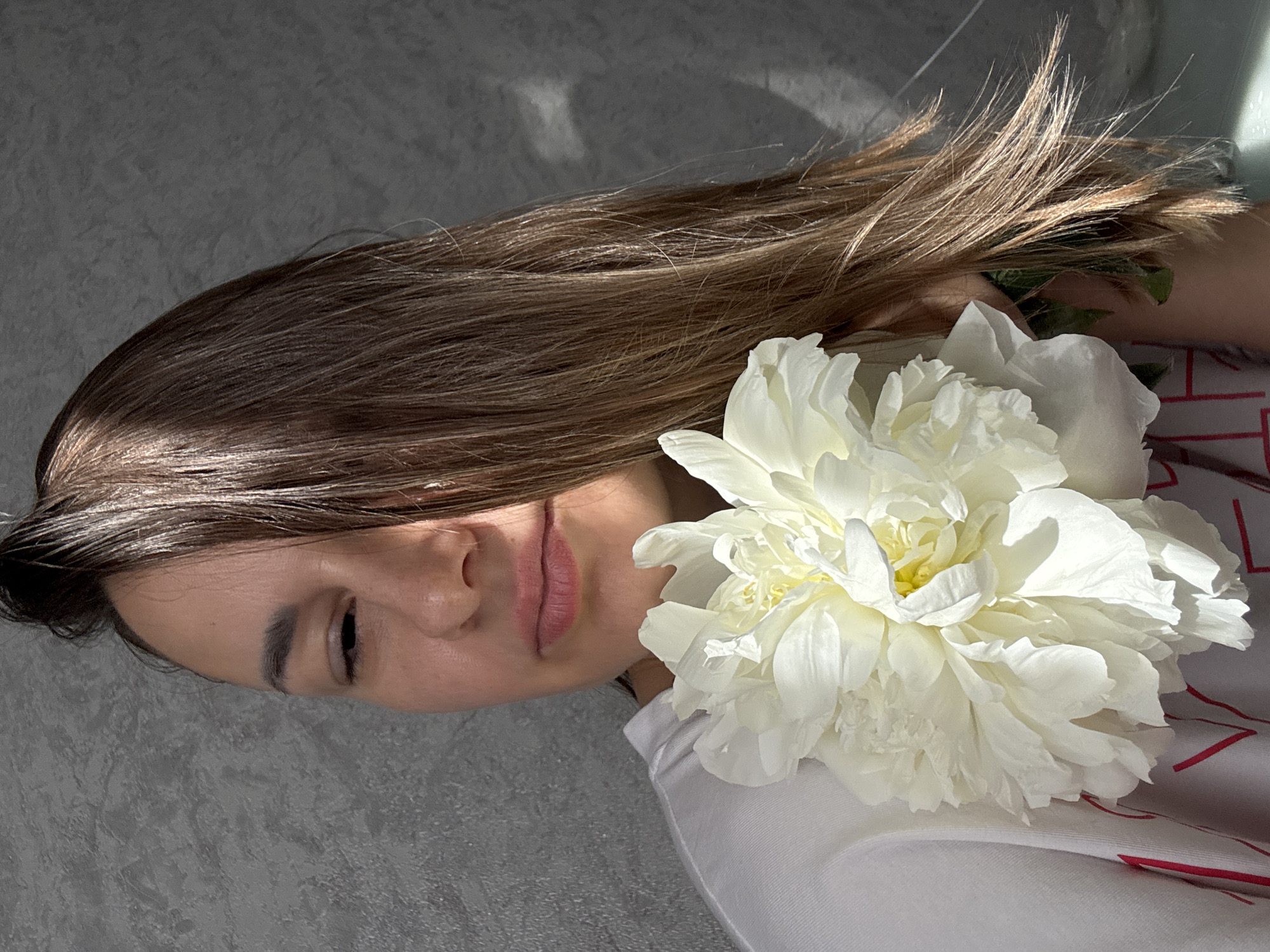 This close-up portrait features a young woman lying on a textured grey surface, her head oriented to the viewer's left. Her eyes are gently closed or mostly closed, and her lips are softly parted, conveying a serene expression. Her long, light brown hair spreads out behind her head, illuminated by strong, directional sunlight that creates defined highlights and shadows across her hair and the background.
A large, pristine white peony flower, in full bloom with a subtle yellow center, is placed prominently beside her head and rests on her upper chest, partially nestled within her hair. The woman is wearing a white garment, possibly a t-shirt, which has faint red linear patterns visible on the fabric near her right shoulder.
The backdrop is a consistent, textured grey surface, suggesting a wall or floor. The lighting indicates bright daylight, casting diagonal shadows that add depth to the composition. The overall mood is peaceful and contemplative. The context provided for the image is Sochi, Russia. No legible text is visible within the frame.