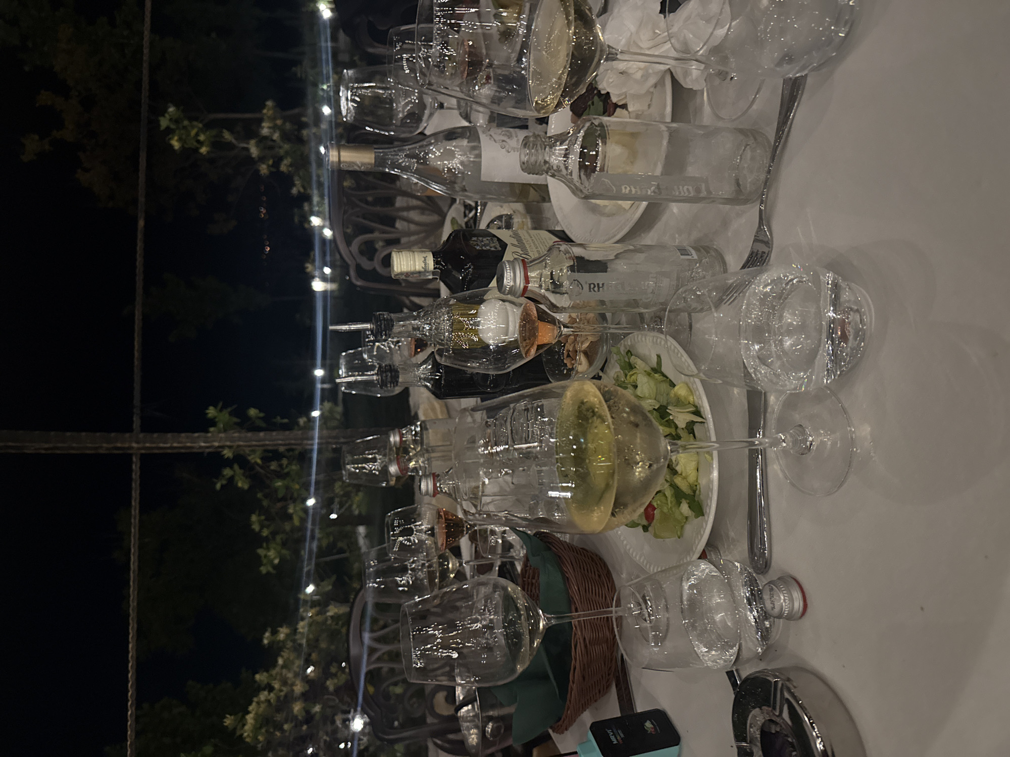 The image depicts a table setting outdoors, likely in a restaurant or event space in Kocherinovo, Bulgaria, given the mention of the city. It is nighttime, with string lights visible in the background among trees.
The table is covered with a white cloth and is cluttered with used glassware, including wine glasses and empty bottles of alcohol. Some plates and silverware are also present, along with a salad on a plate. A basket with a green cloth is visible near the bottom of the frame. A mobile phone is near the edge of the table.