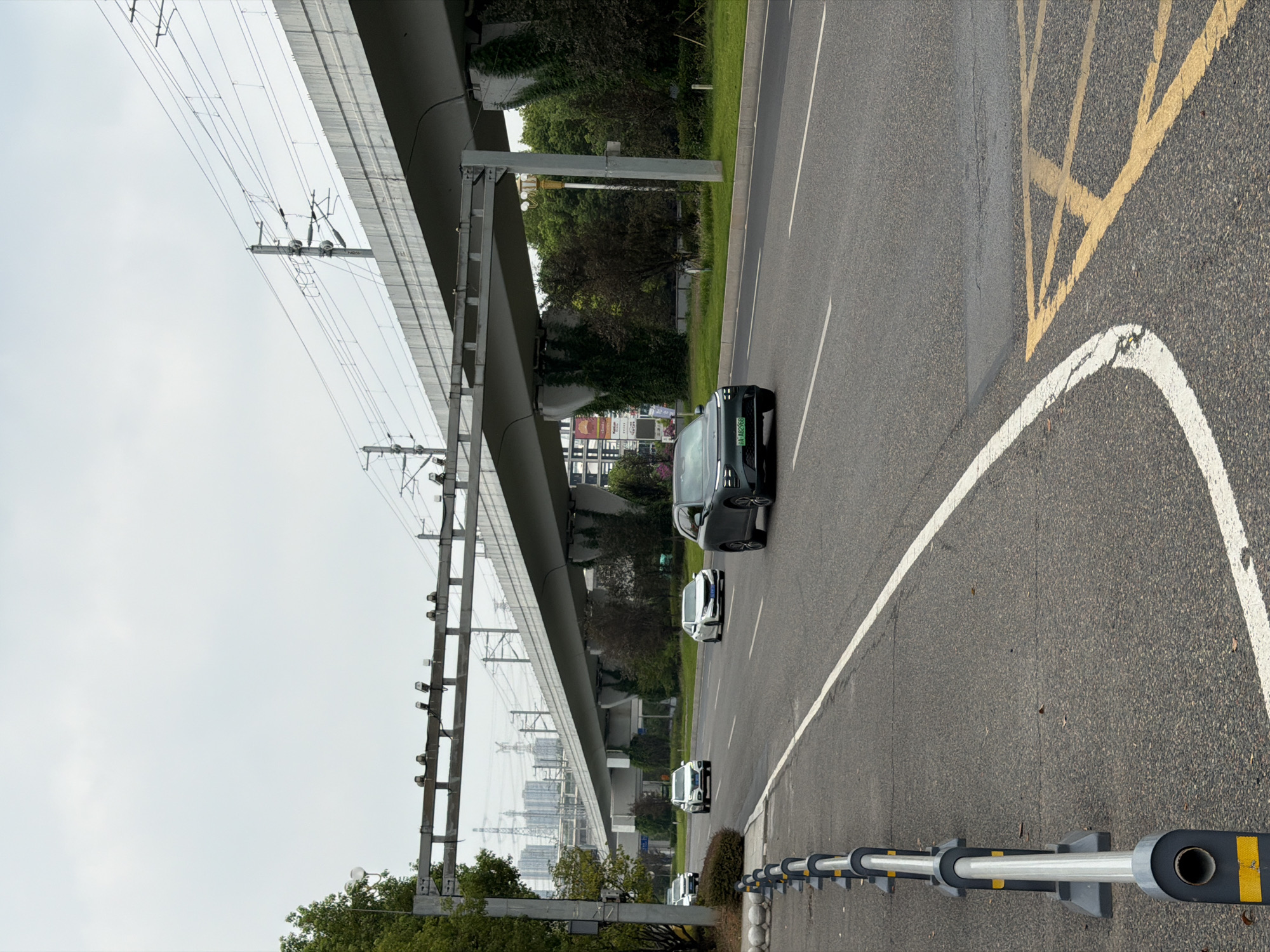 The image captures a street scene in Jiangjin, China, on an overcast day. The foreground shows a portion of an asphalt road with white lane markings and yellow directional arrows painted on its surface. A grey and yellow metal barrier runs along the left edge of the road.
In the midground, several cars are visible on the road. A dark grey electric car is prominently featured in the right lane, with its license plate reading "A·482968". Other cars, including a white sedan and a white SUV, are driving in the same direction. Beyond the road, there is a lush green landscape with trees.
Dominating the background is an elevated concrete structure, likely an overpass or a section of an elevated railway line, supported by substantial pillars. Power lines and poles are strung above and alongside this structure. In the distance, hazy city buildings and industrial structures are visible, hinting at the urban environment of Jiangjin. The sky is cloudy, suggesting a subdued lighting condition. There are no people directly visible in the image.
