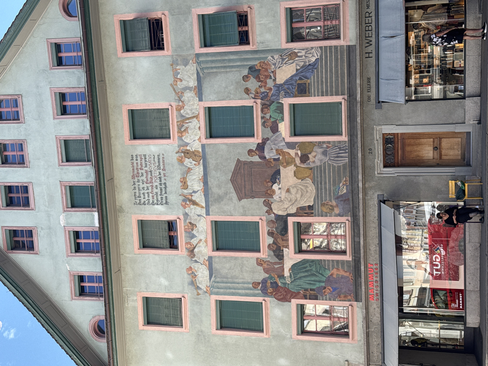 The image shows the facade of a building in Lucerne, Switzerland, featuring a large mural depicting a historical scene with numerous figures. The mural is painted on the upper stories of the building, which has pale green walls and pink-trimmed windows. The scene in the mural includes a group of people gathered around a table, seemingly engaged in a communal meal or gathering. Some figures are playing musical instruments, while others are seated or standing. Below the mural, there are shopfronts at street level. One shop has a large window displaying merchandise, and a person is seated on a yellow chair outside another shop. The building has multiple floors with windows, some of which have green shutters. The architecture suggests an older European city. The mural is signed "EDUARD RENGGLI 1925" at the bottom right. The location is further identified by the signage "MAMMUT SWISS 1862" above one of the shopfronts.