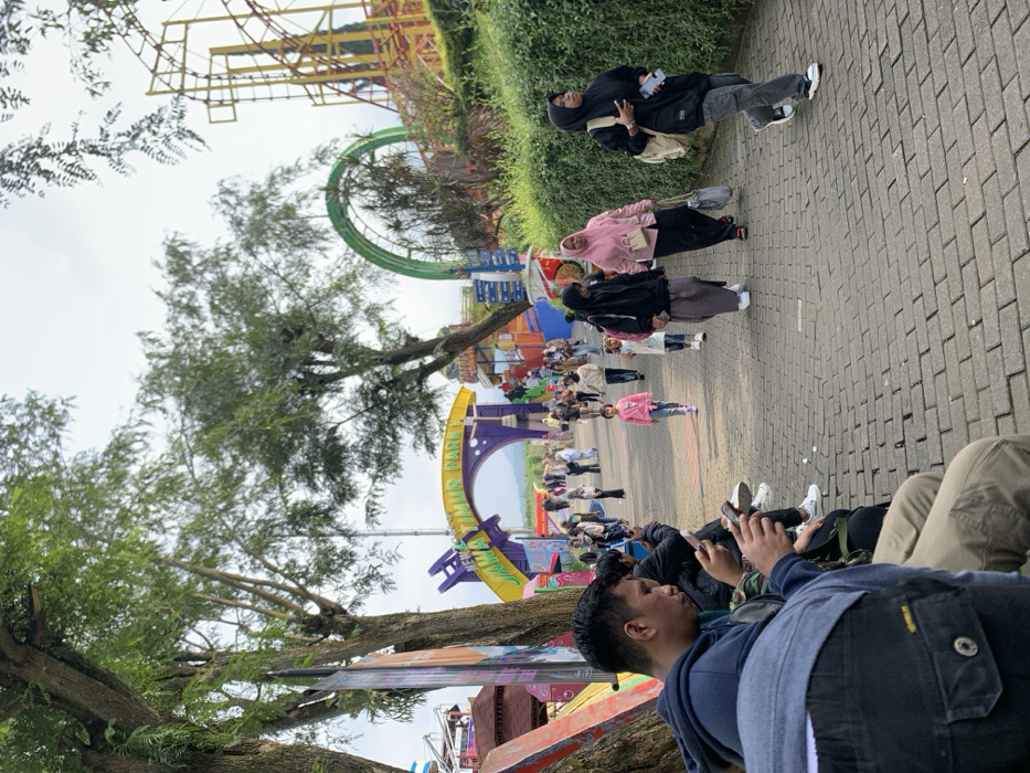 The image shows a group of people walking on a brick pathway at Jawa Timur Park 1 in Batu, Indonesia, located at latitude: -7.885, longitude: 112.524. The park entrance is visible in the background, adorned with colorful arches and signs. A young girl wearing a pink shirt walks towards the entrance, while several other people walk in the opposite direction. A group of four people are sitting on a bench along the pathway, all looking down at their mobile phones. To the right of the pathway is a green bush, and beyond that, a roller coaster track and a large Ferris wheel can be seen in the distance. The sky is overcast and gray.