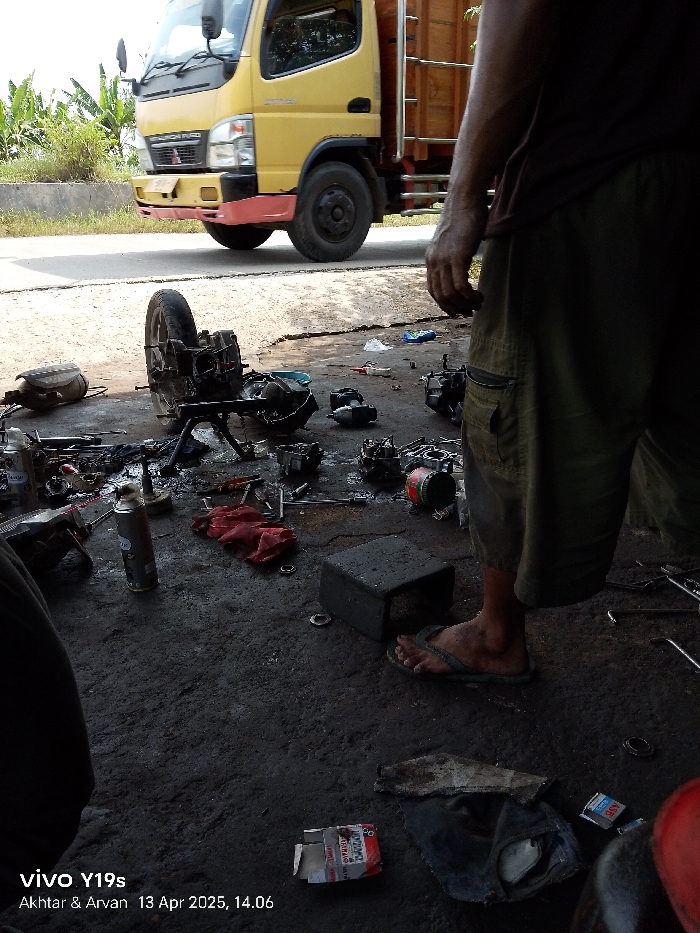 The image shows a scene in Kecamatan Haurgeulis, Indonesia (latitude: -6.414, longitude: 107.953). A person is standing next to a motorcycle that has been partially disassembled. The motorcycle is lying on its side, with its engine parts scattered around. The person is wearing sandals and shorts. They are looking down at the motorcycle. There are several tools and pieces of the motorcycle lying on the ground. In the background, a yellow truck is parked. The scene is set in a parking lot, with a dirt surface and a small amount of vegetation in the background. The photo was taken on April 13th, 2023, at 2:06 PM. The person appears to be inspecting the engine. There is a red cloth, a black box, and a spray can on the ground next to the motorcycle. The person is holding a cigarette in their right hand. The image was taken with a VIVO Y19s camera.