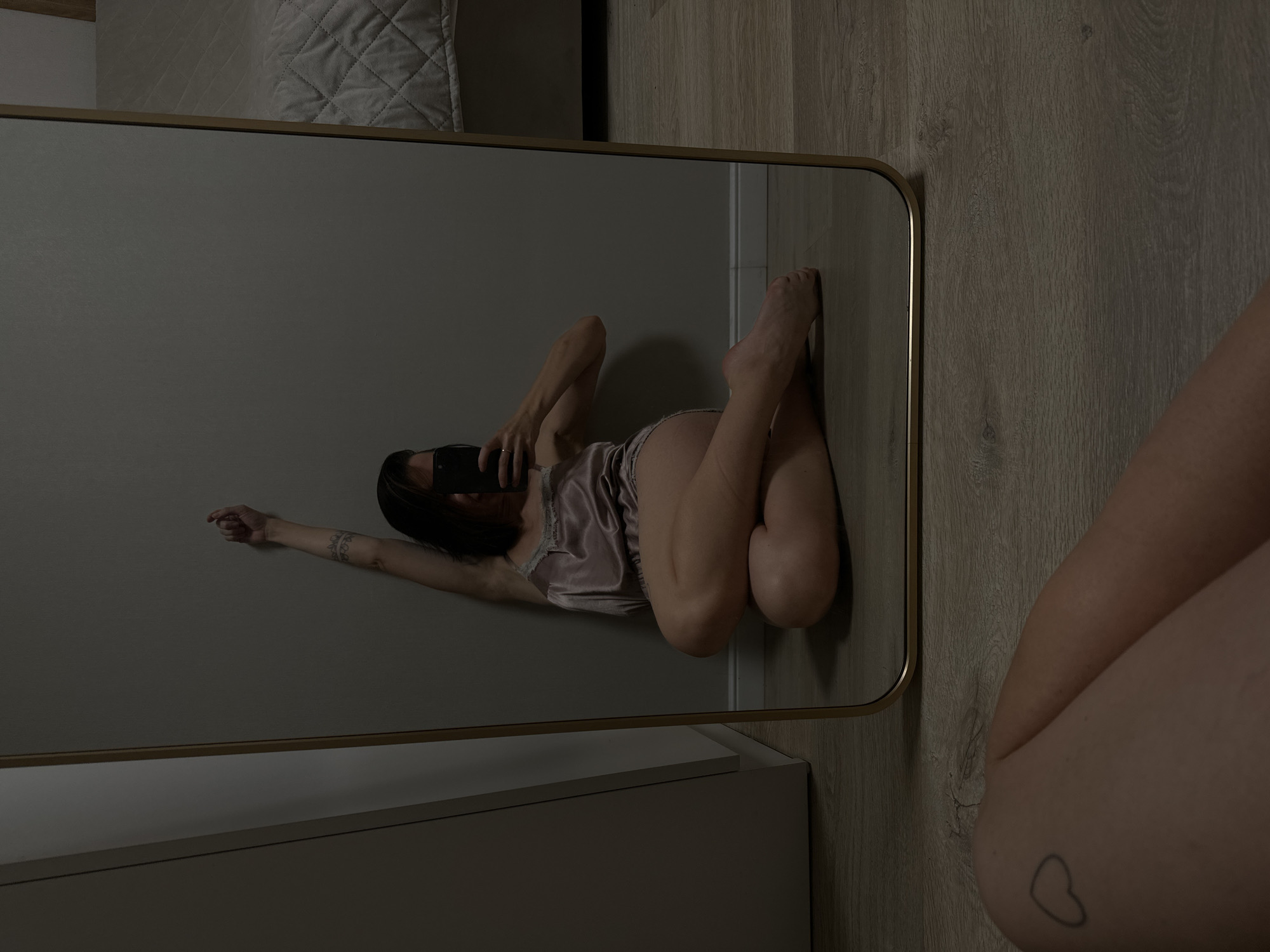 A woman is sitting on the floor in front of a mirror, taking a selfie with her phone. She is wearing a light pink slip dress and has her right arm raised. Her legs are crossed, and her feet are bare. The mirror reflects her image, as well as a portion of a bed and a wall. The floor is made of wood. The woman has a tattoo of a heart on her left thigh. The setting appears to be a bedroom. The image is dark and moody, with the primary light source coming from an unseen source to the left of the frame. There is no information in the image to suggest a location in Votkinsk, Russia.
