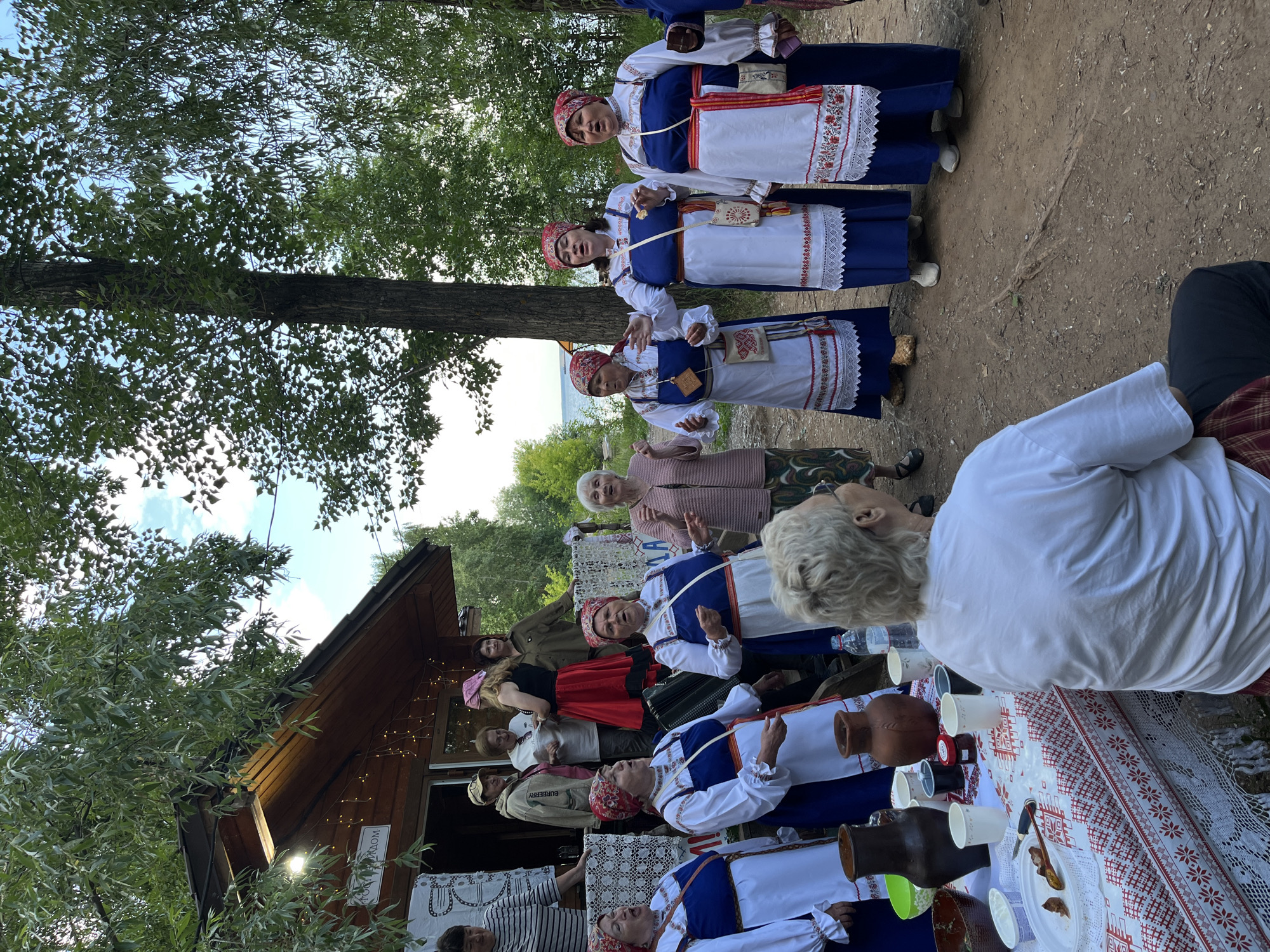 The image depicts an outdoor gathering near a wooden building, likely a cafe or shop, with fairy lights strung across its eaves. Several individuals are present, with a group of women in traditional folk costumes taking center stage. These women, adorned in blue dresses with white embroidered tops and red headscarves, appear to be singing or performing. One woman among them is playing an accordion. Another older woman in a pink cardigan stands to the right, her arms raised. In the foreground, a woman in a white t-shirt is seated, facing away from the camera, with a table covered in a red and white patterned tablecloth set before her. On the table are various items including pottery jugs, bowls, cups, and jars of jam. The environment suggests a natural setting with trees and a glimpse of water in the background, possibly a river or lake. This scene is consistent with events held in natural landscapes near Chaikovsky, Russia, such as those organized at tourist sites along the Kama River.