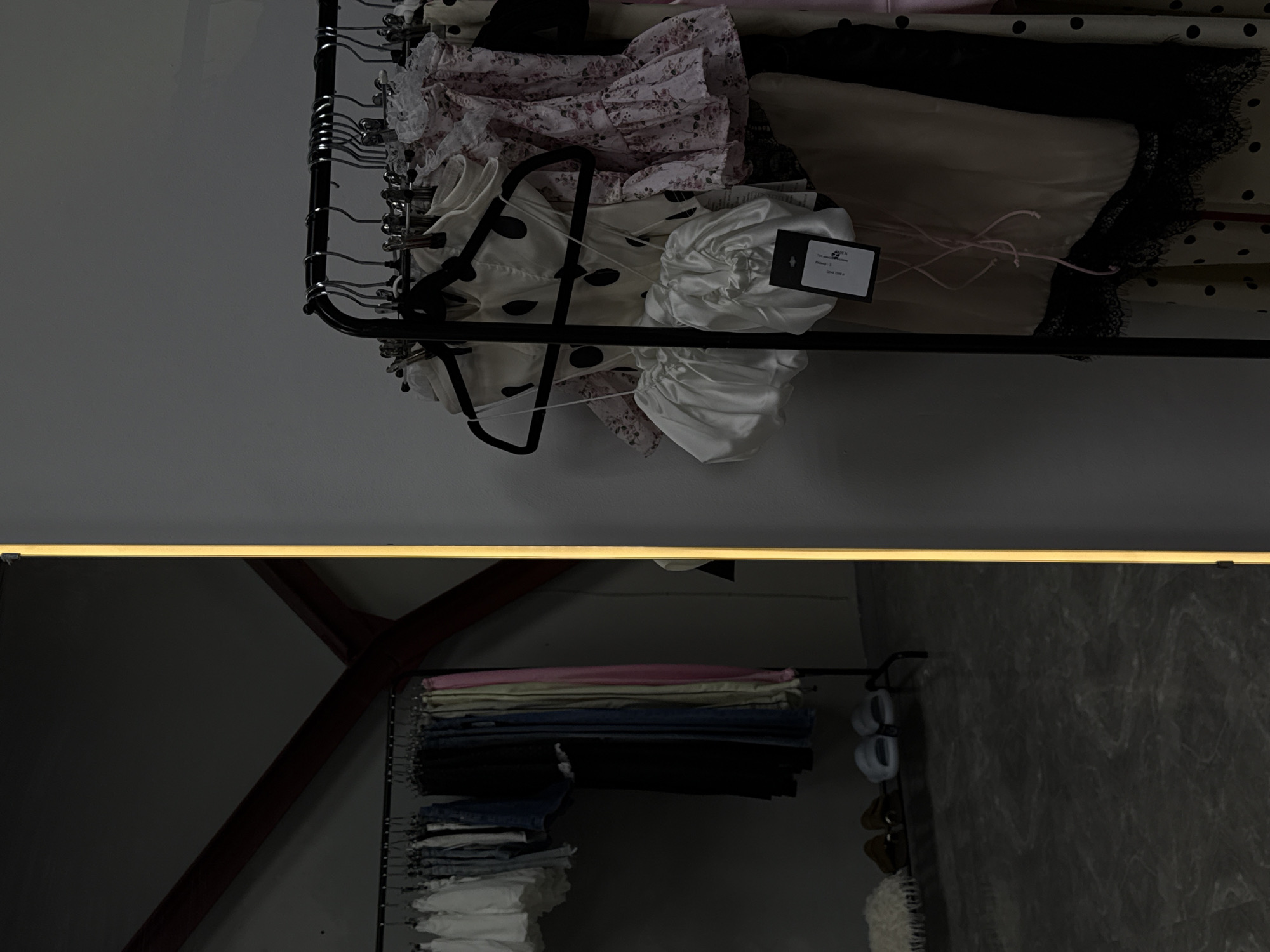 This media file shows a close-up of clothing items displayed on a rack. The clothing appears to be a collection of delicate, possibly feminine garments, including dresses or tops with floral patterns, polka dots, and lace trim. Some items are white and feature ruching or gathering details. A tag is visible on one of the white garments, with some text printed on it.
Below the rack of delicate clothing, the reflection shows another rack with folded clothing, possibly jeans and t-shirts, in various colors such as blue, black, pink, and white. There are also a pair of light blue slides and a pair of brown sandals visible in the reflection, suggesting a personal closet or boutique setting. The overall lighting is dim, with a bright, diffused light source at the top of the frame, casting a warm glow. The background is a plain gray wall, with some dark wooden beams visible in the reflection, suggesting an interior space. The scene evokes a sense of organized storage for clothing.
The visible text on the tag is: "ART 432 N. / Printed 2 / Price: 1990 р.". Given the location context of Novosibirsk, Russia, "р." likely stands for rubles.