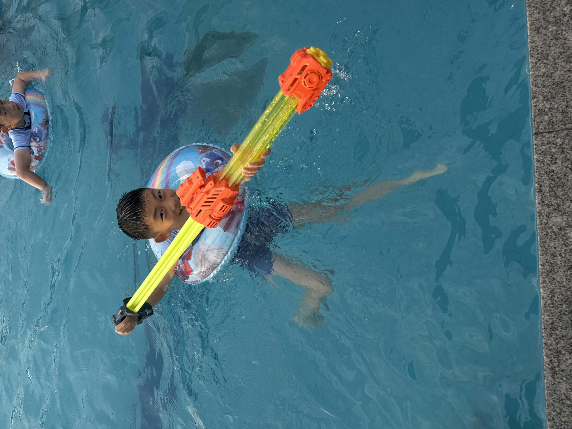 An overhead shot captures two young boys enjoying a sunny day in a swimming pool. The boy in the foreground, wearing a colorful swim ring, holds an orange and yellow water gun. His dark hair is wet, and he looks directly at the camera with a playful expression. He is wearing blue swim trunks. The second boy is in the background, also in a swim ring and appears to be floating or swimming. The water is a clear blue, with visible ripples and reflections. The edge of the pool, made of speckled gray stone, is visible on the right side of the frame, indicating an outdoor setting. The lighting suggests it is daytime, likely with good weather for swimming. The overall scene is one of childhood fun and summer activity. No text is visible in the image.