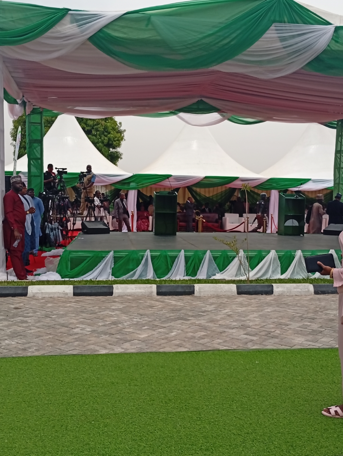 This image captures an outdoor event setting, likely a formal gathering or ceremony in Unguwan Sanusi, Nigeria. The central focus is an elevated stage draped in green and white fabric, possibly mirroring the Nigerian national colors. Overhead, a canopy is fashioned from flowing white, green, and soft reddish-pink draped fabrics, providing shade.
On the stage, two prominent green structures, appearing to be podiums or speaker stands, are visible. Several people are on or around the stage; some are seated, potentially attendees or dignitaries, while others are standing. To the left of the stage, a cluster of professional video cameras on tripods, manned by individuals, indicates significant media coverage or recording of the event. Among the people visible are men in traditional Nigerian attire, such as a man in a red robe and white cap, and others in suits, suggesting a mixed formal dress code.
In the background, large white marquee tents are set up, extending the event space, with some greenery and trees visible beyond them. The foreground features a neatly laid paved area with interlocking bricks, separated by a curb with black and white stripes from a stretch of artificial green turf. The overall lighting suggests it is daytime, possibly overcast, creating soft, diffused light. There is no visible text in the image.