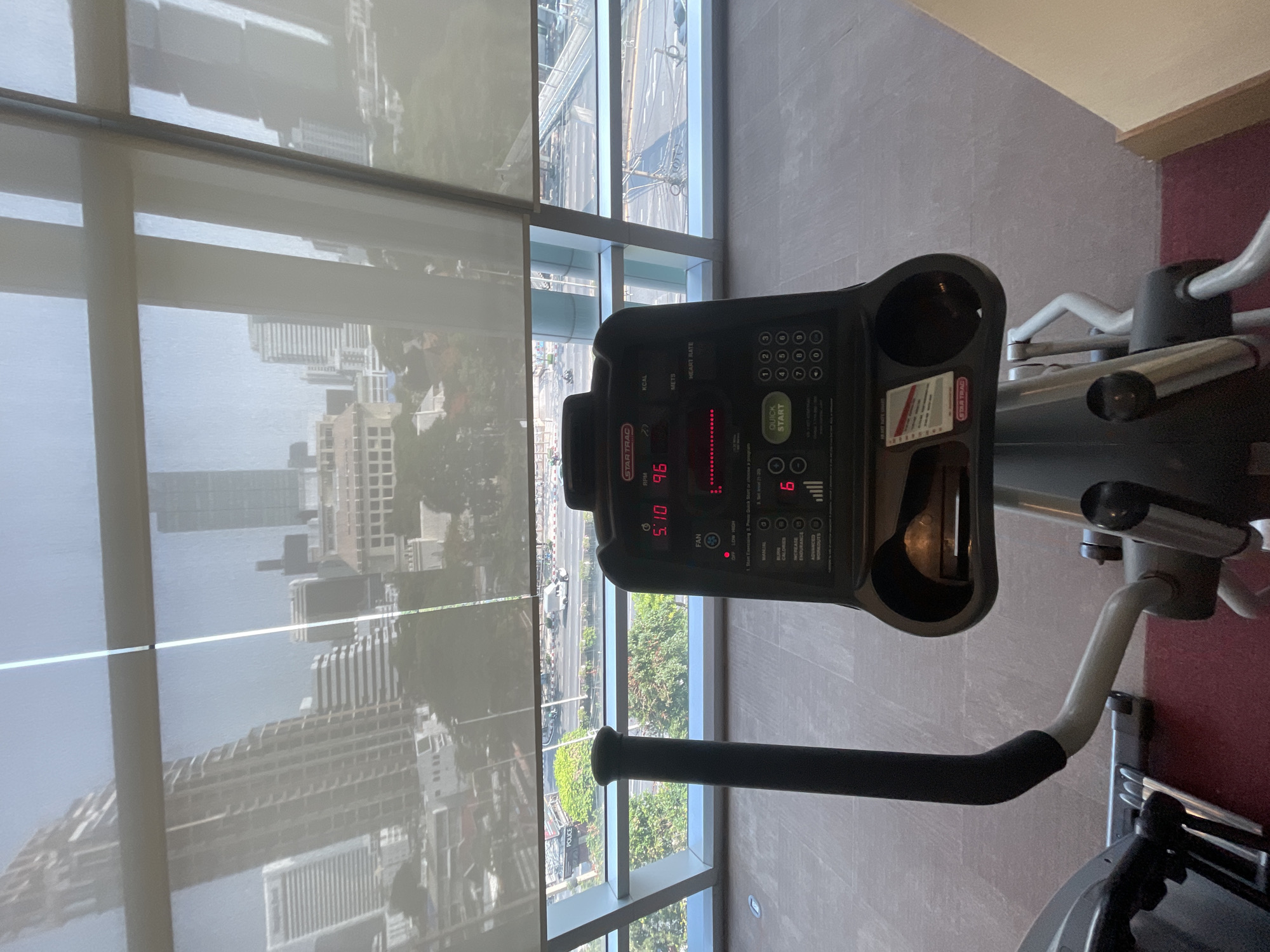 The image shows an indoor fitness area with an elliptical machine placed next to a large window, overlooking a city. The view is captured from a slightly elevated perspective, looking down onto the exercise equipment and the urban landscape beyond.
In the foreground, the console of a "STAR TRAC" brand elliptical machine is prominent. Its digital display shows "5:10" (likely time elapsed) and "96" (likely RPM). Other visible text on the console includes "KCAL," "METS," "HEART RATE," "FAN," "QUICK START," "INCREASE ENDURANCE," and "ADVANCED WORKOUTS," along with a numeric keypad. Two cup holders are integrated into the console.
Through the expansive window, which features horizontal blinds or shades, a bright daytime view of Bangkok, Thailand, is visible. The cityscape includes numerous high-rise buildings and a multi-lane road. On the street below, a light-colored vehicle with the word "POLICE" clearly marked on its side can be seen. The overall setting suggests a modern gym or fitness facility within a building, offering a panoramic view of the surrounding city. No people are visible in the scene.