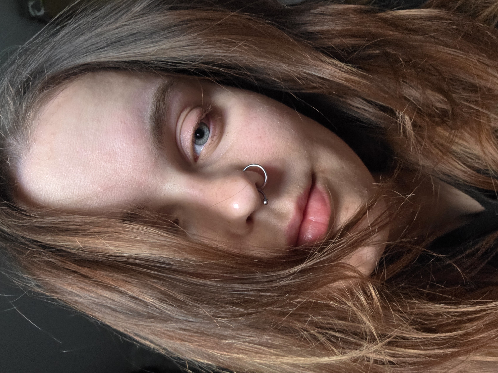 Close-up portrait of a person with fair skin, light blue eyes, and reddish-brown hair. The person has a silver hoop nose piercing. The hair is disheveled and partially obscures the face. The background is dark and indistinct. Location: Unknown, Unknown.