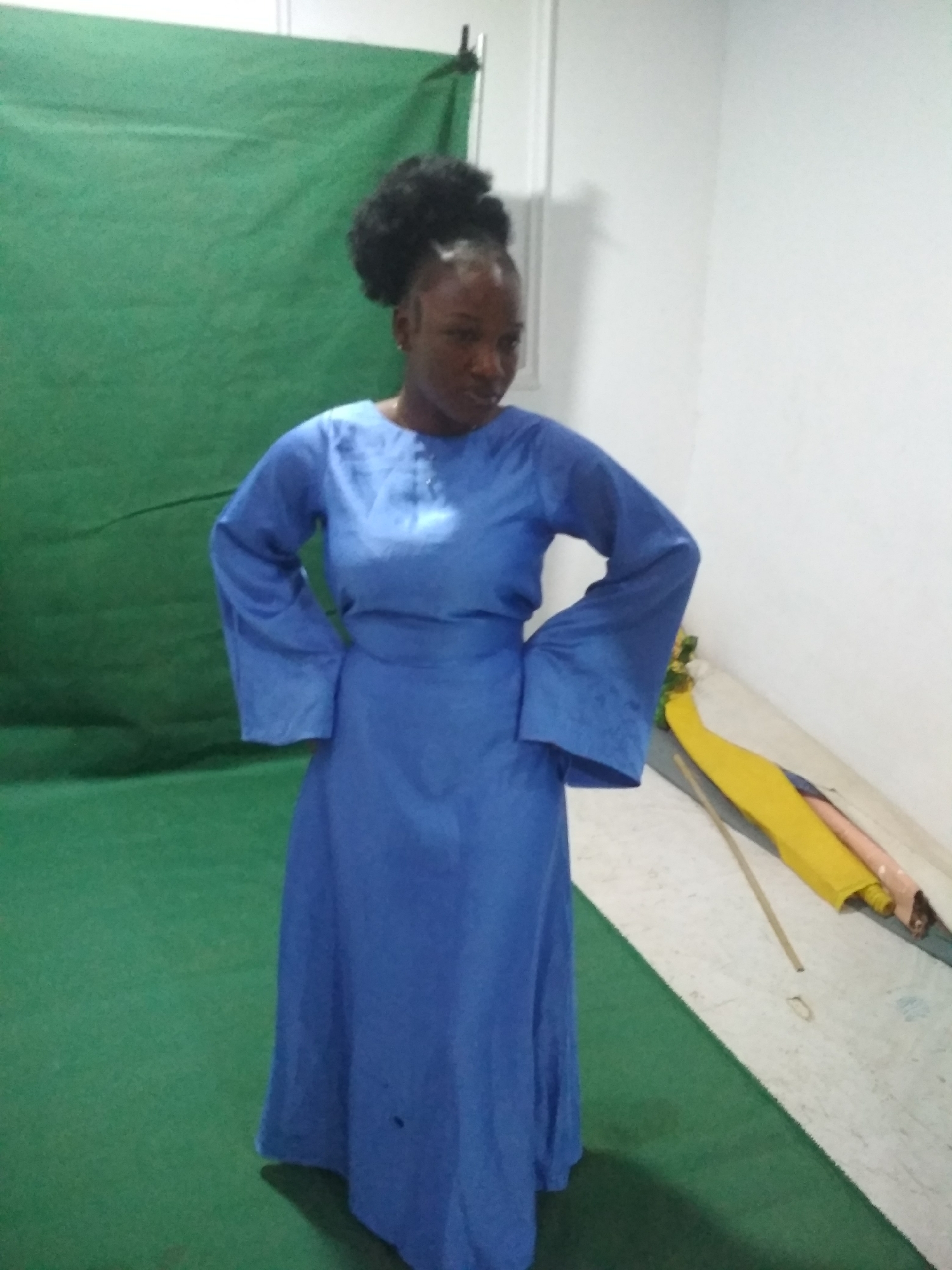 A young woman stands in a room. She has dark skin and dark hair styled in a bun. She is wearing a long, blue dress with bell sleeves and stands with her hands on her hips.
The background is a green screen, with a white wall visible on the right side of the frame. There are some objects leaning against the wall, including a yellow fabric and a tan or peach colored object.
The location is Unknown, Unknown.