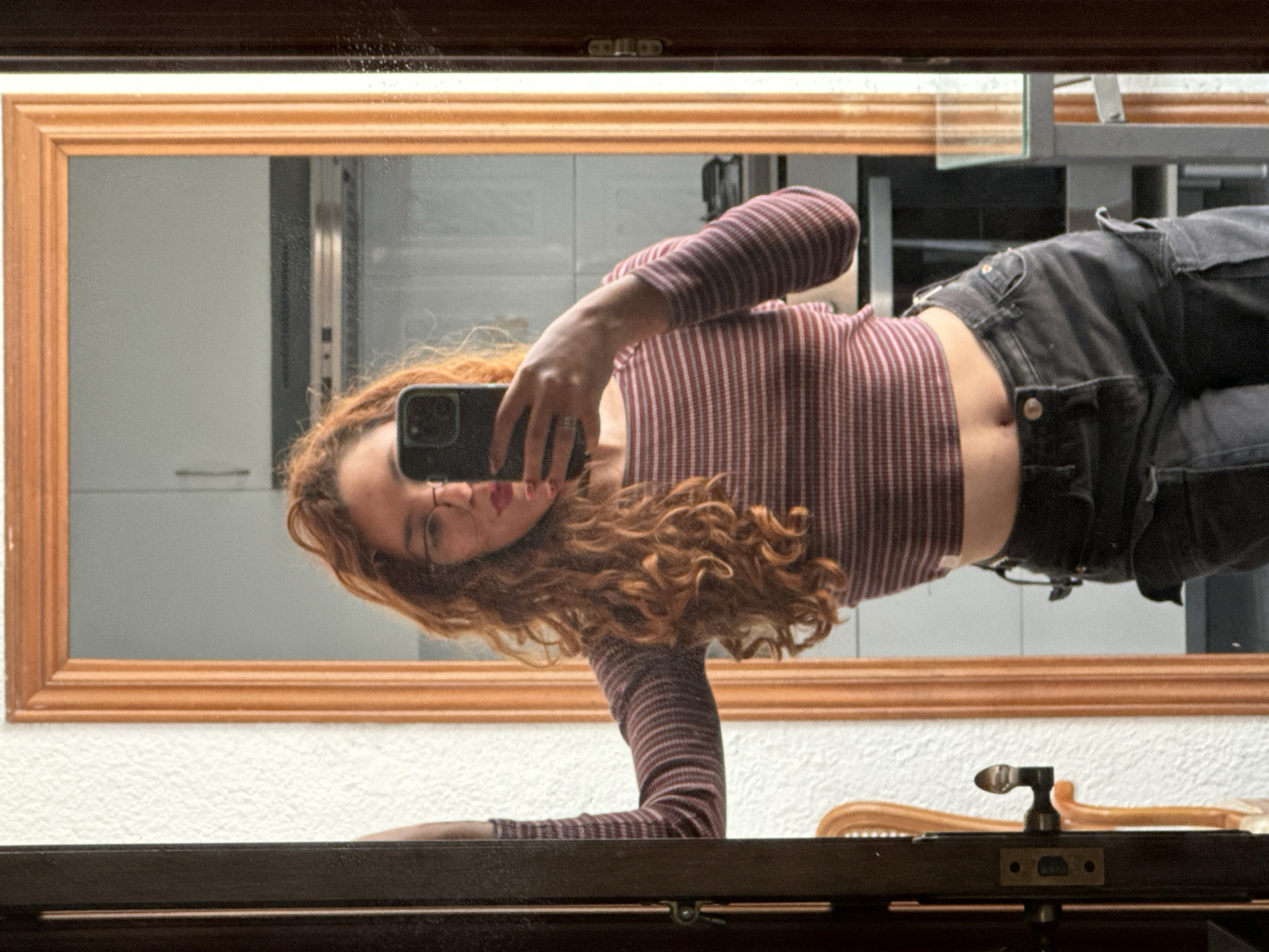 A mirror selfie captures a young person with long, curly, reddish-brown hair and glasses, holding a black smartphone. The image is rotated 90 degrees clockwise, showing the person's reflection lying horizontally across the frame with their head to the left. She is wearing a reddish-brown and white striped long-sleeved crop top, revealing her midriff, and dark cargo pants.
Her left arm (from her perspective) is bent, holding the smartphone to capture her reflection, while her right arm is bent and rests on a dark brown surface in the foreground. The selfie is taken in a mirror with a light brown wooden frame. The background reflected in the mirror behind the person appears to be an indoor setting with light-colored walls and indistinct architectural features or furniture. Outside the mirror frame, a textured white wall is visible. On the dark brown surface in the lower right, a metallic door handle or latch mechanism and part of a curved wooden chair back are present. The lighting suggests it is daytime. The scene is set in Barcelona, Spain. There is no visible text in the image.