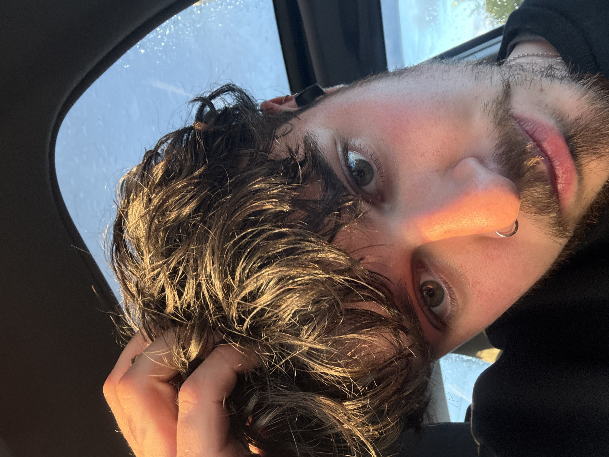 A close-up, tilted selfie shows a man with dark, wet hair in a car. The sunlight reflects brightly on his hair. He has a visible nose ring and a short beard. His right hand is touching his hair. The car's sunroof and window are visible, with water droplets on the glass. The city and location are unknown.
