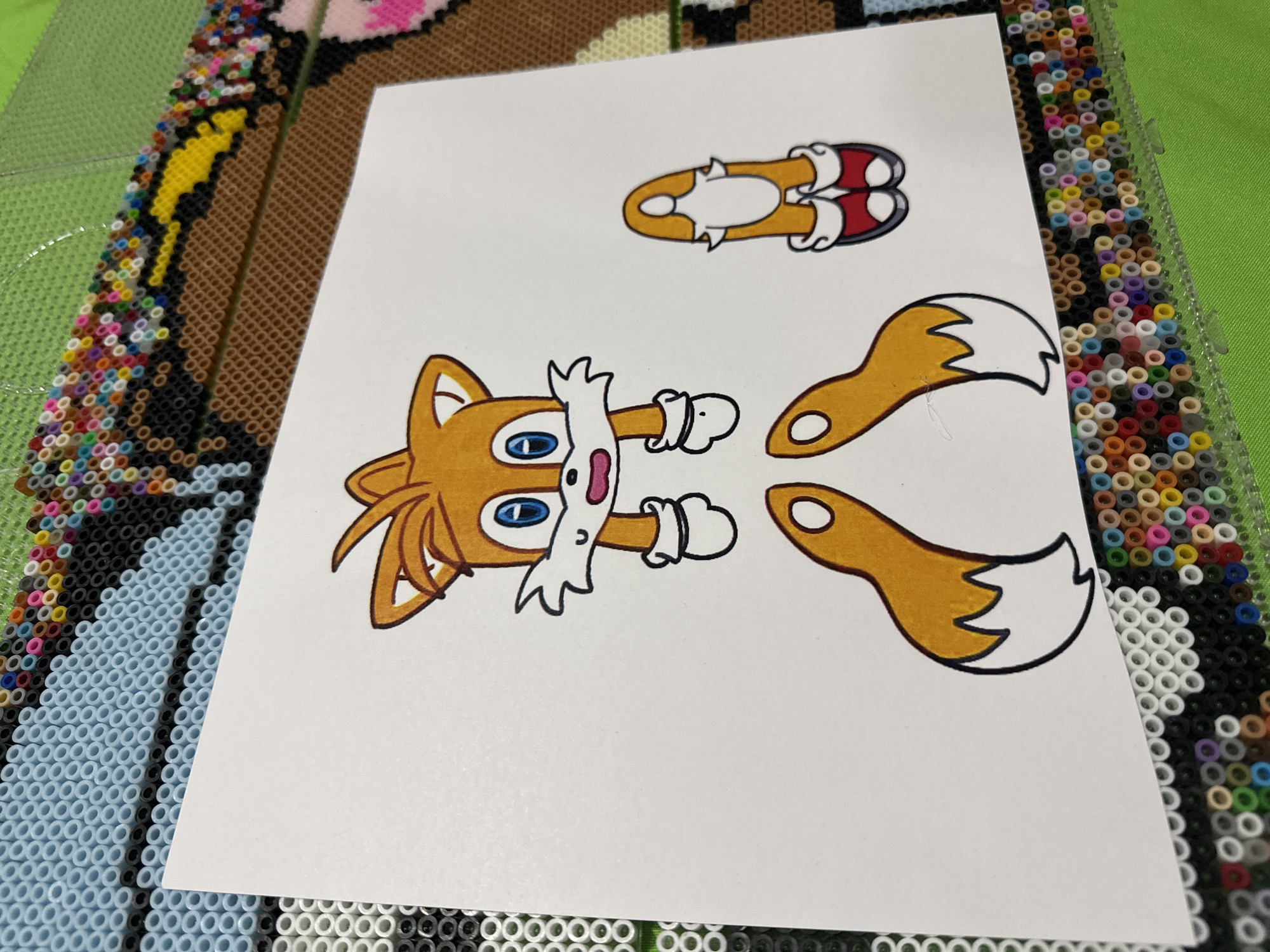 The image presents a white sheet of paper with cartoon drawings of Tails from the Sonic the Hedgehog franchise. The drawings depict Tails' head and torso, his running shoes, and his two tails. The paper is placed on top of a grid of Perler beads, featuring a variety of colors. A portion of a Perler bead image featuring light blue, black, and pink can be seen. In the upper left corner, a section of yellow and brown beads is visible. The background suggests the scene is likely a crafting or hobby area. The location details (city of Puebla, Mexico) are not visually apparent in the image.