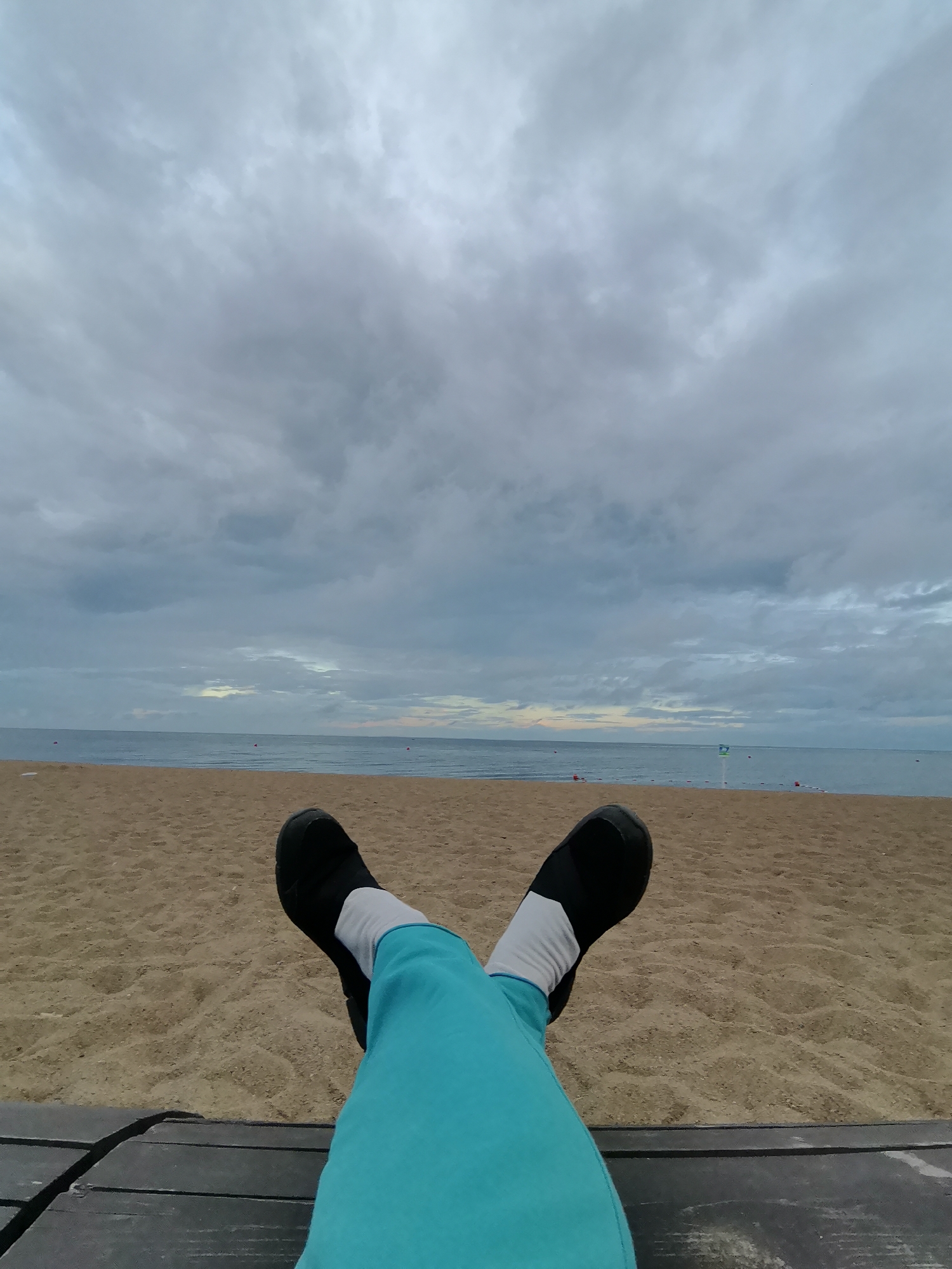 The image shows a person's legs and feet from a first-person perspective, resting on a wooden platform at a beach. The person is wearing blue pants and black slip-on shoes with white socks. The beach is sandy, and the ocean stretches out to the horizon under a cloudy sky. In the distance, there are small buoys in the water. The scene appears to be a quiet and overcast day at a beach in Zelenogorsk, Russia.