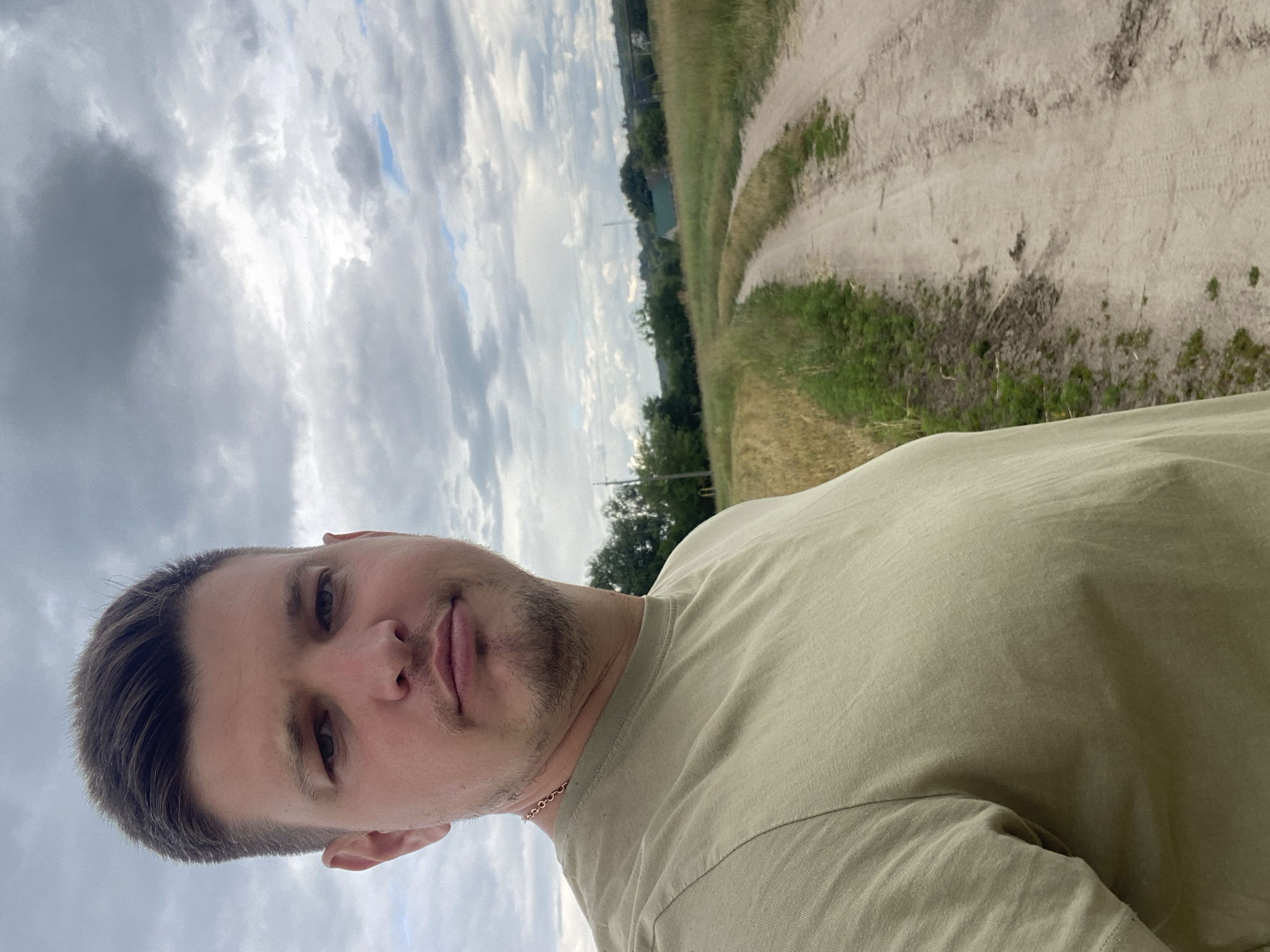 A man stands close to the camera, facing slightly left, with a dirt road stretching into the distance behind him. He has short, dark hair and is wearing a light green t-shirt. A thin gold chain is visible around his neck. The road is flanked by fields of dry grass and green vegetation. In the background, trees and a few buildings can be seen, suggesting a rural or semi-rural setting. The sky is overcast with clouds. The image appears to be taken in Yablunivka, Ukraine.
