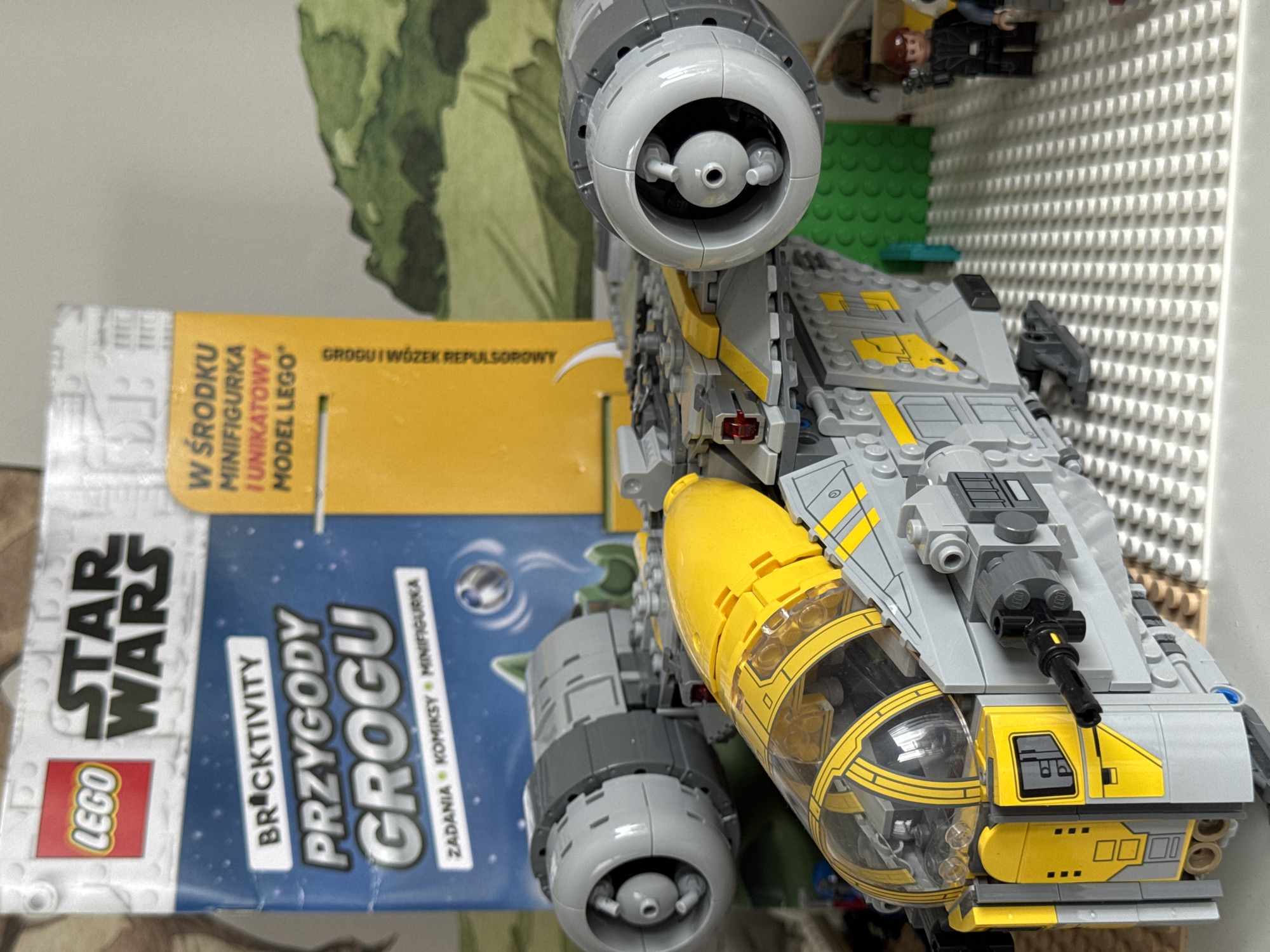 In an indoor setting, a detailed Lego Star Wars model spaceship is the central focus. The spaceship, primarily gray and yellow with a transparent cockpit, is positioned on a white Lego baseplate. Behind the spaceship, a Star Wars Lego activity book is visible, featuring the title "PRZYGODY GROGU" and "LEGO STAR WARS." The book is partially obscured by the spaceship. To the right of the spaceship, several Lego minifigures are present, including one in black attire holding a weapon, and another in brown attire. The background includes a green Lego surface and a suggestion of a landscape, possibly a printed backdrop or another Lego creation. The lighting suggests the image was taken indoors with natural or artificial light. The location is Unknown, Unknown.