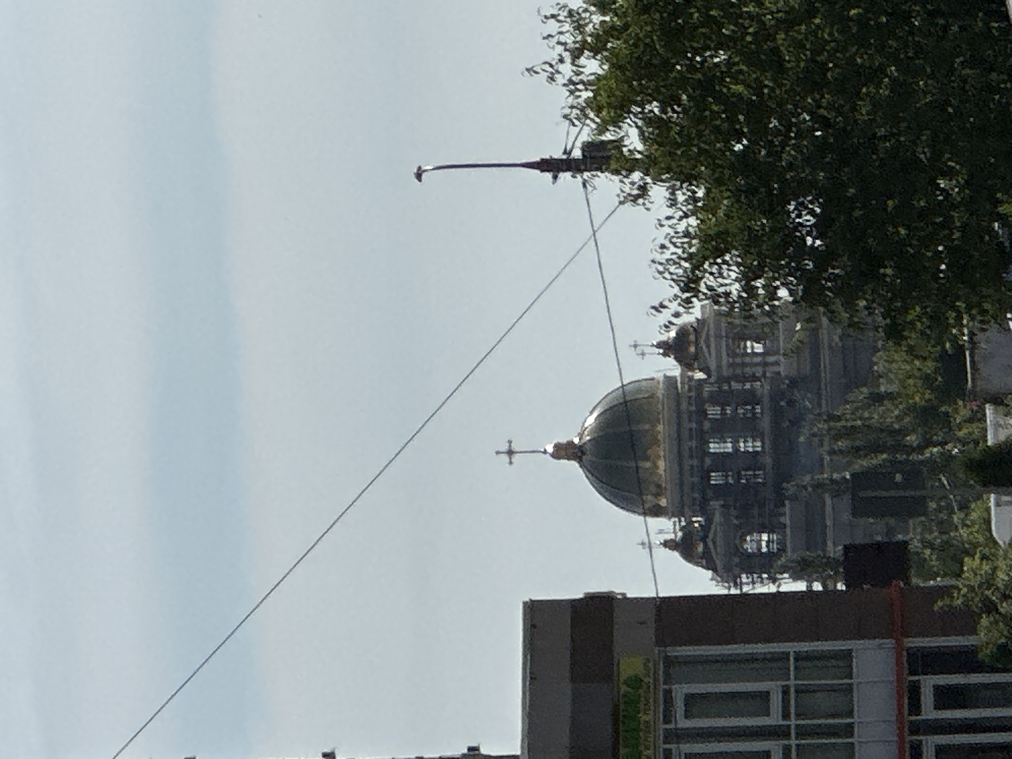 The image captures a view of a large, domed building, likely a church or cathedral, with a golden dome topped by a cross. The building appears to be undergoing construction or renovation, with scaffolding visible on its facade. In the foreground, a portion of a modern building with windows is visible on the left, and dense green trees partially obscure the view on the right. A tall lamppost stands to the right of the domed structure. Power lines crisscross the sky above. The overall scene suggests an urban environment with architectural landmarks. The image is taken on a clear day with a light blue sky. Based on the architectural style and the presence of a cathedral, this is likely the Spassky Cathedral in Podolsk, Russia.