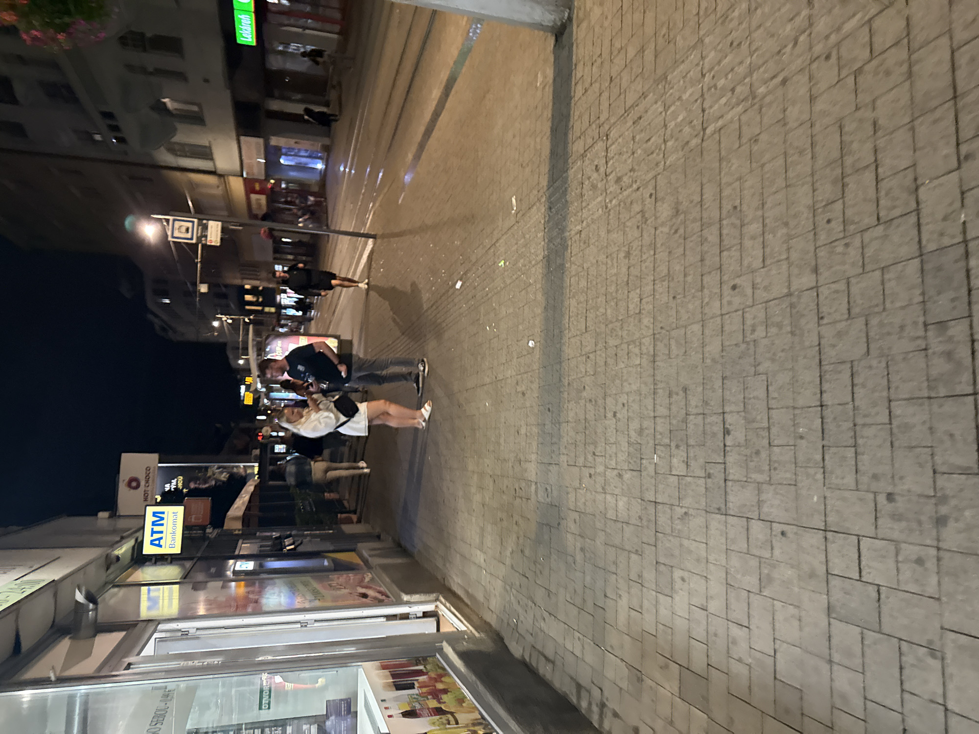 The image depicts a street scene at night in Bratislava, Slovakia. The perspective is from a low angle, looking down a paved walkway that borders a street with tram tracks. On the left, a storefront with a glass display is visible. Further down the street, a couple is standing and talking. The man is wearing a dark t-shirt and shorts, while the woman is in a white dress and white sandals. In the background, other pedestrians are walking, and buildings line the street, with various signs and lights illuminated. Some litter is scattered on the pavement and street. A sign for "ATM Bankomat" is prominently displayed on the left side of the image, indicating a banking service. The overall atmosphere is that of a typical urban environment at night, with artificial lighting creating reflections on the wet pavement.