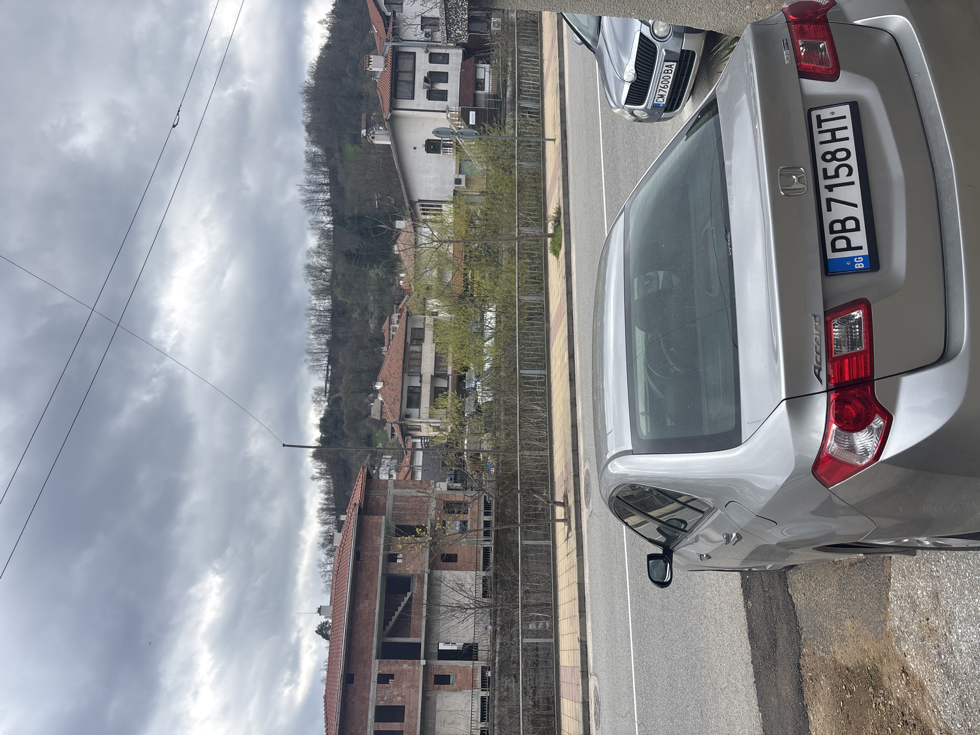 The image captures a street scene in Zlatograd, Bulgaria. A silver Honda Accord, bearing the Bulgarian license plate PB7158HT, is parked on the right side of the frame. Adjacent to it, partially visible, is a silver Skoda with license plate CM7600BA. The street is lined with a yellow and beige patterned sidewalk. Across the street, there is a building under construction, appearing to be made of brick. Behind this, are houses and trees extending up a hillside. The sky is overcast with a cloudy appearance. Overhead power lines are visible in the upper left portion of the image. The scene is still and there are no people present.