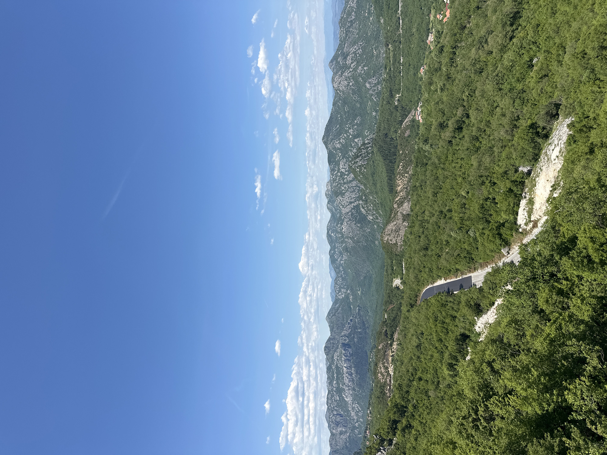 The image is a landscape view from Bukovik, Montenegro. The sky is a clear blue with scattered white clouds. A series of rocky mountains stretch across the center, with vegetation on their slopes. A winding road is visible on the right side, bordered by green trees. The landscape is rugged and mountainous.