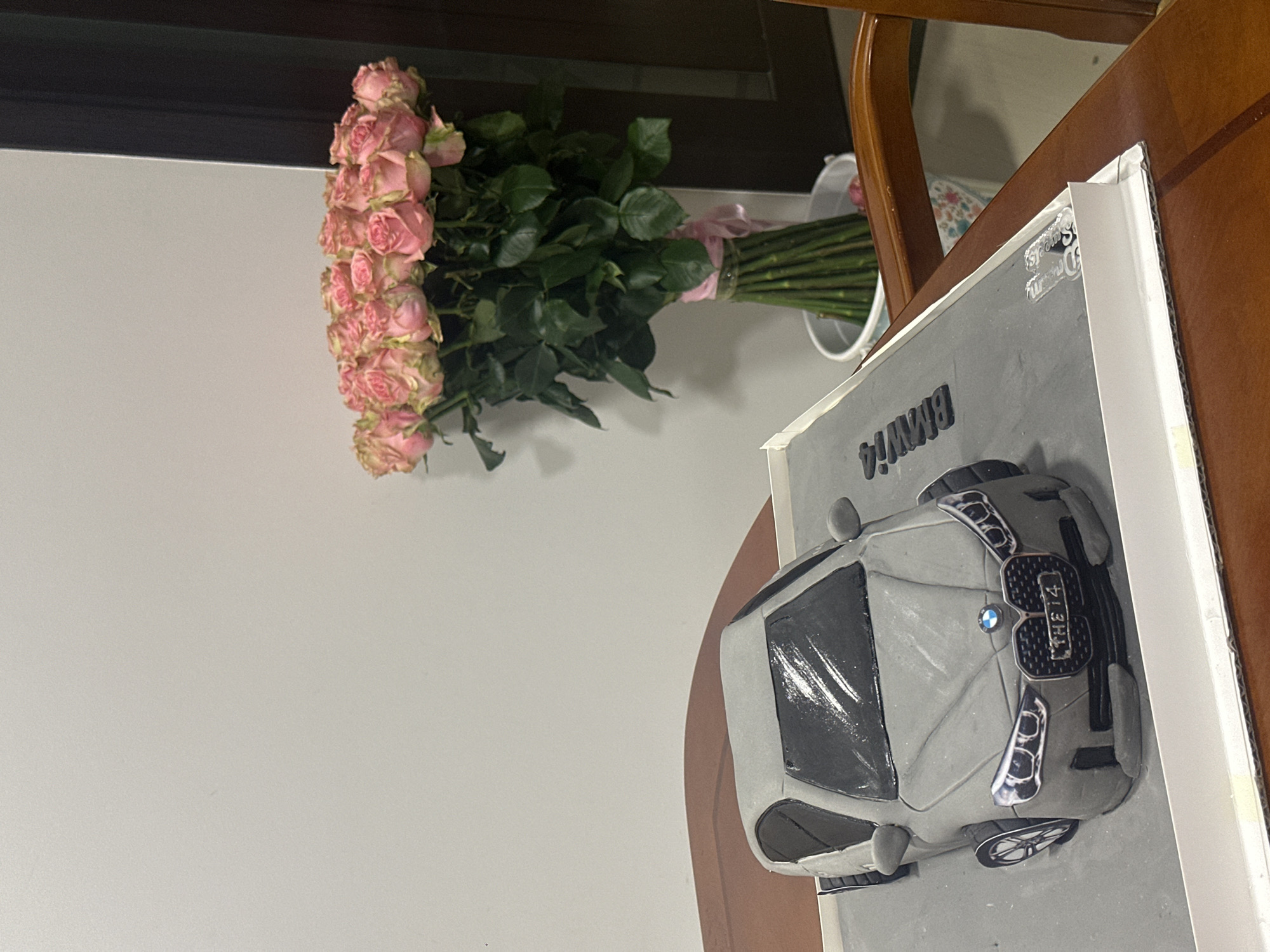 A grey BMW car cake sits on a table in front of a bouquet of pink roses in a white bucket. The cake is detailed, with a license plate that reads "THE i4." The background is a plain wall. The scene appears to be indoors, likely in a domestic setting. No people are visible in the image. The overall impression is one of celebration or a special occasion.