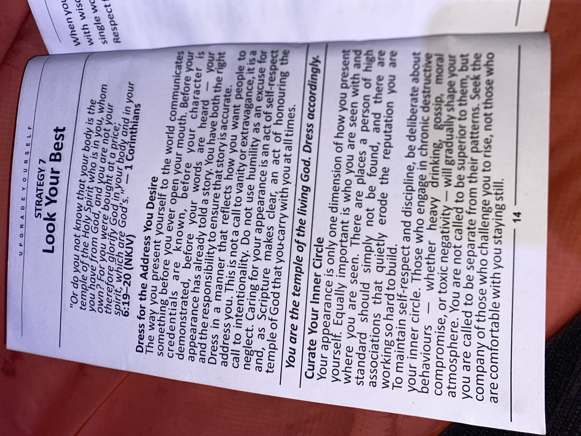 The image shows a close-up of a page from a book or booklet, opened to a section titled "Look Your Best," which is part of a larger theme labeled "Upgrade Yourself." The page contains text about the importance of dressing appropriately and maintaining a positive image, referencing a biblical quote from 1 Corinthians. It also emphasizes the importance of one's inner circle and avoiding negative influences. The page number "14" is visible at the bottom. The image is slightly angled and a person's finger is partially visible at the top of the photo. The location is not Bukuru, Nigeria.