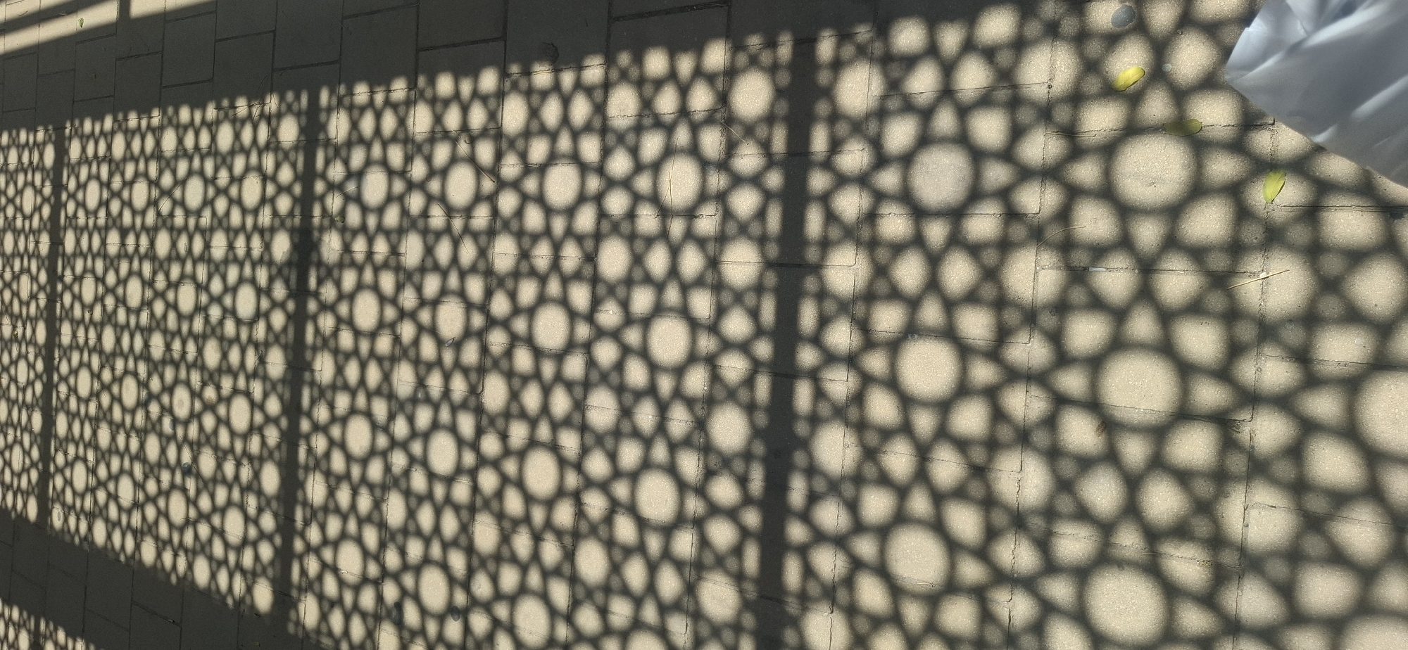 The image depicts a shadow pattern cast by a geometric screen onto a paved surface. The screen exhibits a repeating pattern of stars within circles, creating a tessellated effect. The paving stones appear to be rectangular and laid in a regular grid. Sunlight is strong, casting distinct shadows. The scene is outdoors. The image is taken from a slightly elevated perspective looking down. There are a few scattered leaves on the ground, one of which is bright yellow. In the bottom right corner, a portion of white fabric is visible. The overall impression is one of sunlight and shadow playing on a man-made surface. The location is the city of Yerocham, Israel.