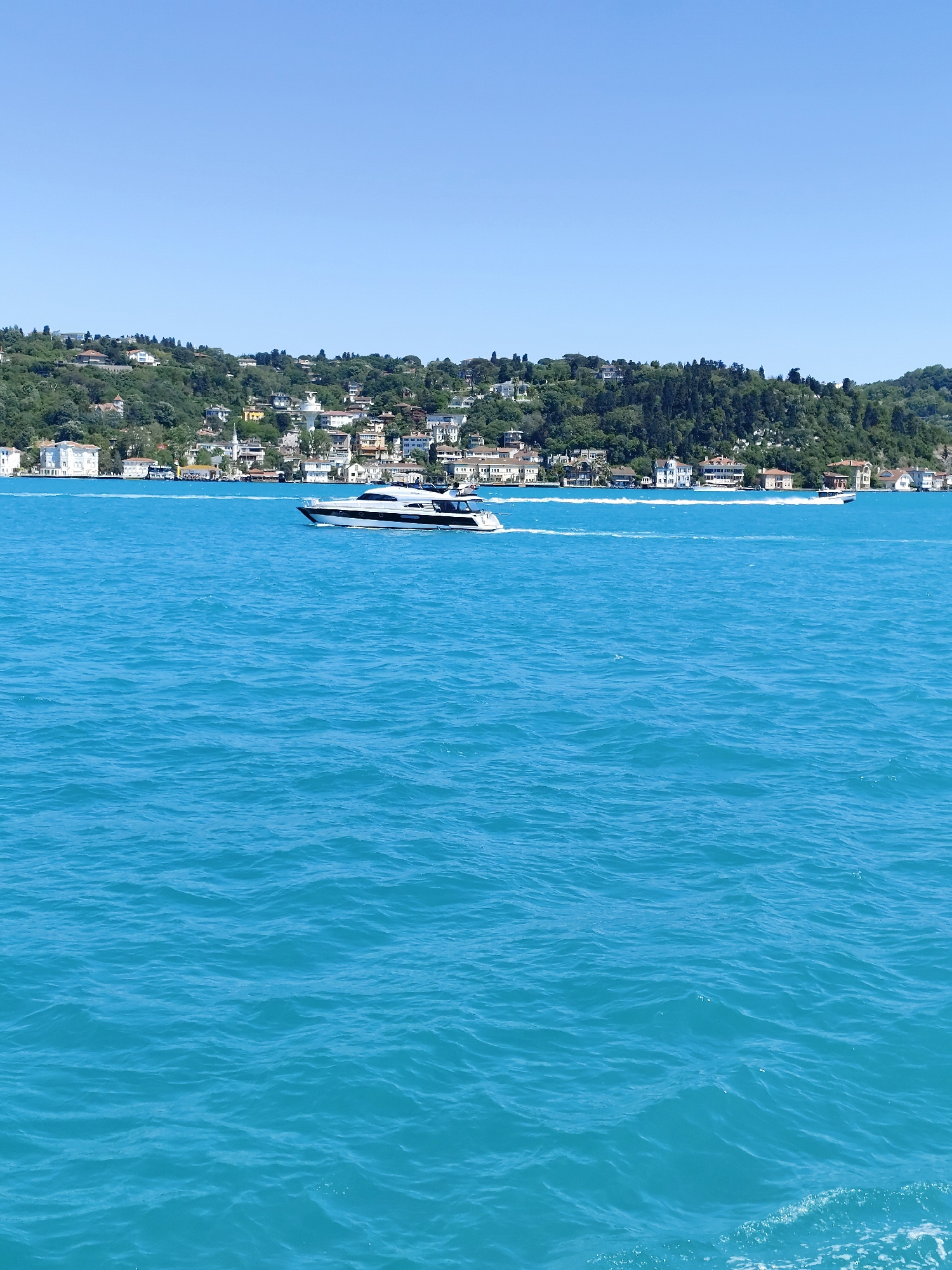 The image shows a view of the Bosphorus in Sarıyer, Turkey. A white yacht is moving across the water, leaving a wake. In the background, a densely populated hillside is visible, covered with trees and houses. The sky is clear and blue. The water is a bright turquoise color.