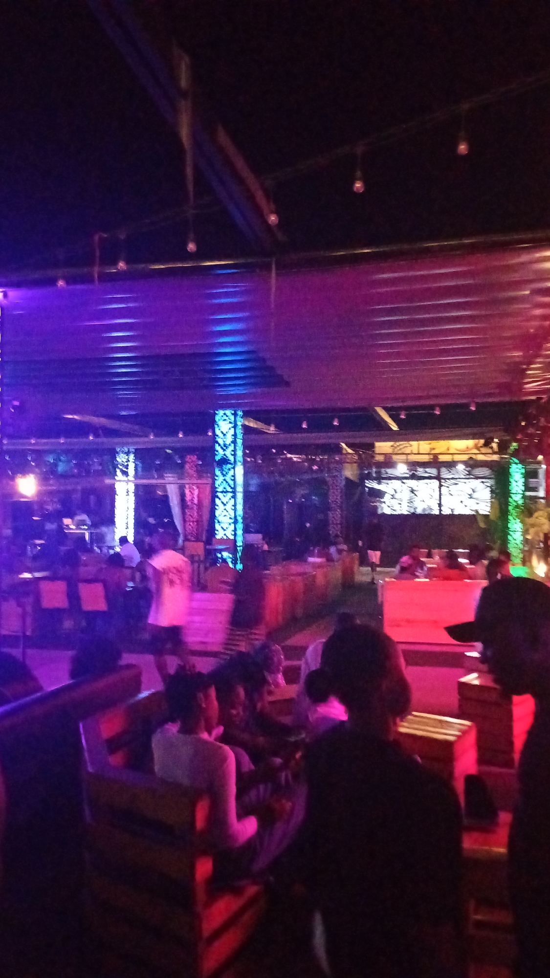 The image depicts an indoor event space with purple and blue lighting. In the foreground, several individuals are seated in wooden benches. Towards the middle ground, there are tables and chairs, some of which are occupied. Prominent architectural features include decorative pillars illuminated with blue light and a slatted ceiling structure. The overall ambiance suggests a social gathering or entertainment venue in Howayi, Nigeria.
