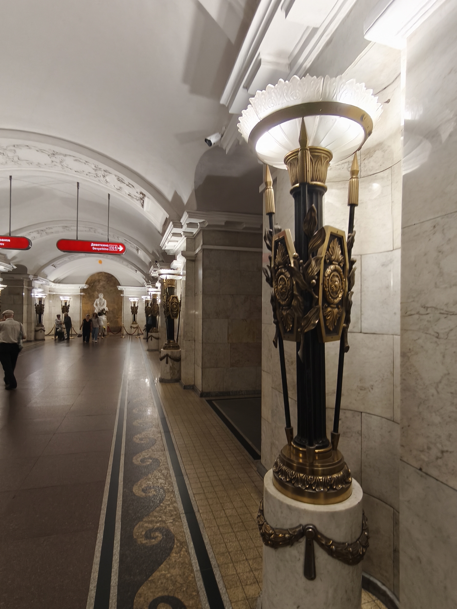 The image depicts the interior of a metro station in Saint Petersburg, Russia, likely Devyatkino station, as indicated by the signage. The scene is characterized by ornate architecture, including a high, arched ceiling with decorative molding, marble-clad walls, and tiled flooring.
In the foreground, a large, ornate lamp post stands prominently. It features a marble base adorned with a golden laurel wreath motif, a dark central column, and a golden decorative element with shield-like shapes and leaf designs. The top of the lamp post has a large, white, lotus-shaped diffuser illuminating the area.
The background reveals a long, wide passageway. On the left side of the passageway, there are red directional signs with white text. People are walking along the passageway, some appearing to be commuters. Further down the passageway, a marble statue of a figure is visible against a curved wall, with decorative railings and more lamp posts lining the sides. The floor is a mix of polished stone and patterned tiles, with a decorative inlay running along the center of the passageway. The overall impression is one of grandeur and historical significance, typical of many Saint Petersburg metro stations.