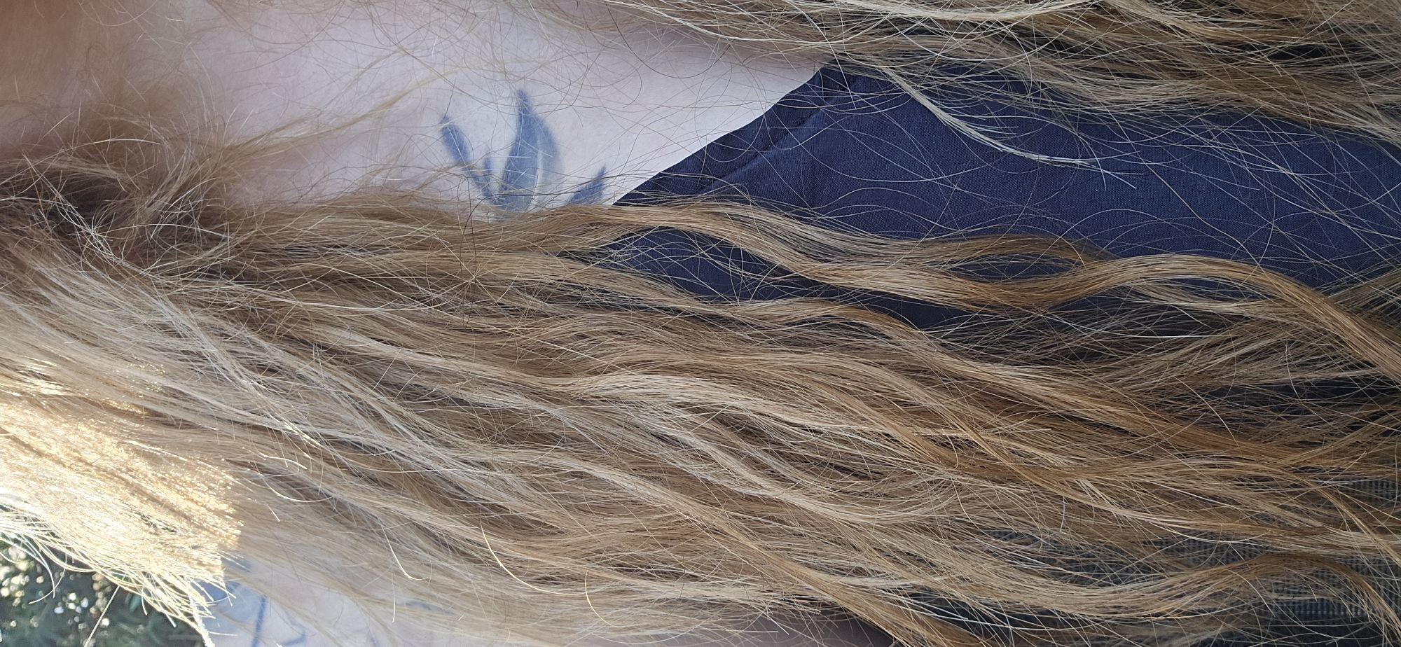 The image is a close-up, vertical shot of a person's head and upper torso, with a dark blue garment visible. Long, wavy, blonde hair dominates the frame, obscuring much of the face. A tattoo resembling stylized wings or tribal markings in a dark blue hue is visible on the person's cheekbone, partially hidden by the hair. The lighting suggests an outdoor setting, with sunlight catching the strands of hair and creating highlights. The background is blurred, with hints of greenery, possibly foliage. The exact location within Radovići, Montenegro, cannot be determined from the image alone.