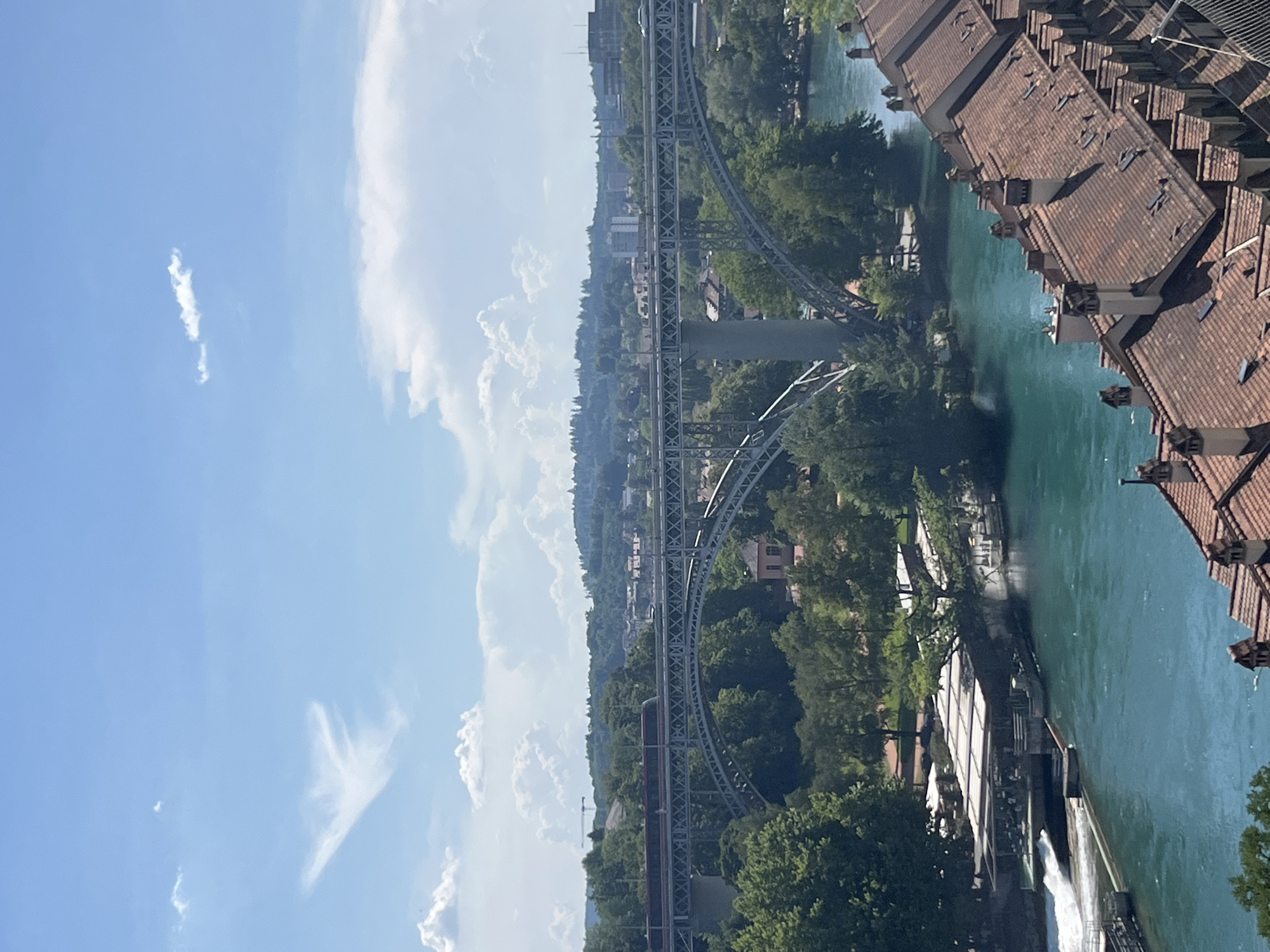 This image captures a wide view of the Aare River in Bern, Switzerland, on a clear day with scattered white clouds. Dominating the mid-ground is a multi-span metal arch bridge, with a train in transit across its top deck. The bridge is supported by large, curved arches and concrete piers.
The river flows from left to right across the frame, its water a vibrant turquoise color. On the left bank, a structure with a white, corrugated roof extends over the water, featuring seating and what appears to be outdoor dining. A small waterfall or spillway is visible to the left of this structure.
The riverbanks are densely populated with lush green trees. In the foreground, a row of traditional Bernese buildings with terracotta tile roofs and numerous chimneys line the right bank, creating a distinct architectural character.
Beyond the bridge, the cityscape of Bern unfolds, with buildings of varying heights nestled among rolling, tree-covered hills that recede into the distance under a bright blue sky. The overall impression is a harmonious blend of urban infrastructure, natural landscape, and historic architecture.