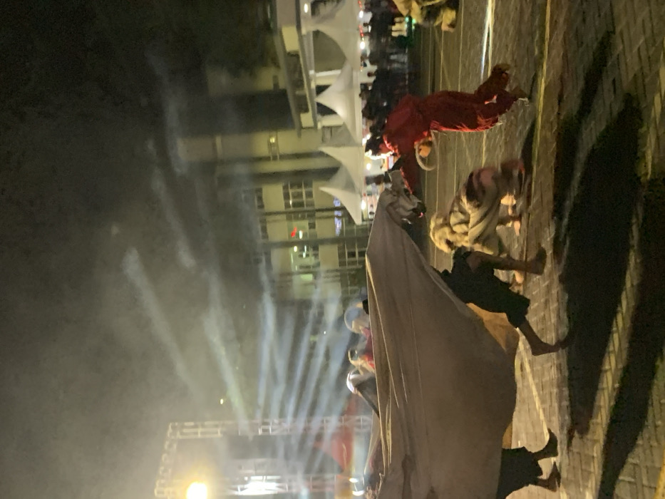 The image shows a nighttime street performance in Kecamatan Batu, Indonesia, a city located at the coordinates -7.867 latitude and 112.514 longitude. The performers are dressed in traditional costumes, with one person wearing a large, white cloth draped over their body, another wearing a black garment, and a third person wearing a red costume. The person in the white cloth is interacting with a person in a tiger costume by holding the tiger's tail. The person in the red costume is running away from the performers. The performance is taking place on a paved street illuminated by streetlights. The background features a building with large windows and multiple white tents. The air is filled with smoke from the performance.