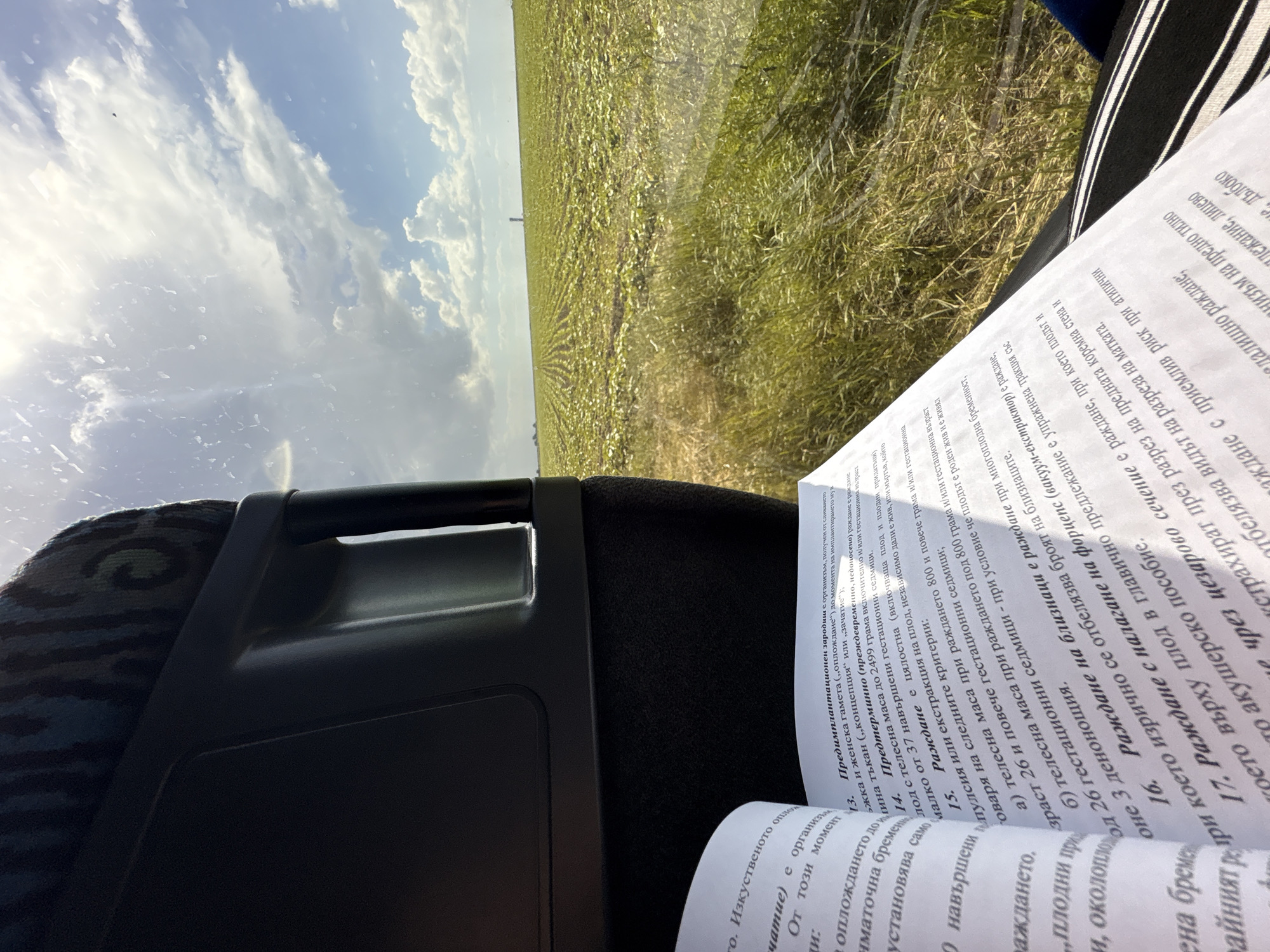 The image appears to be taken from inside a vehicle, possibly a bus or car, offering a view of a field and sky through the window. A book written in Bulgarian is visible on the right side of the frame, possibly on the seat next to the photographer. The location may be Byala Slatina, Bulgaria, considering the text in the book. The scene includes elements of transportation, reading, and a rural landscape. The book contains information on the subject of pregnancy.