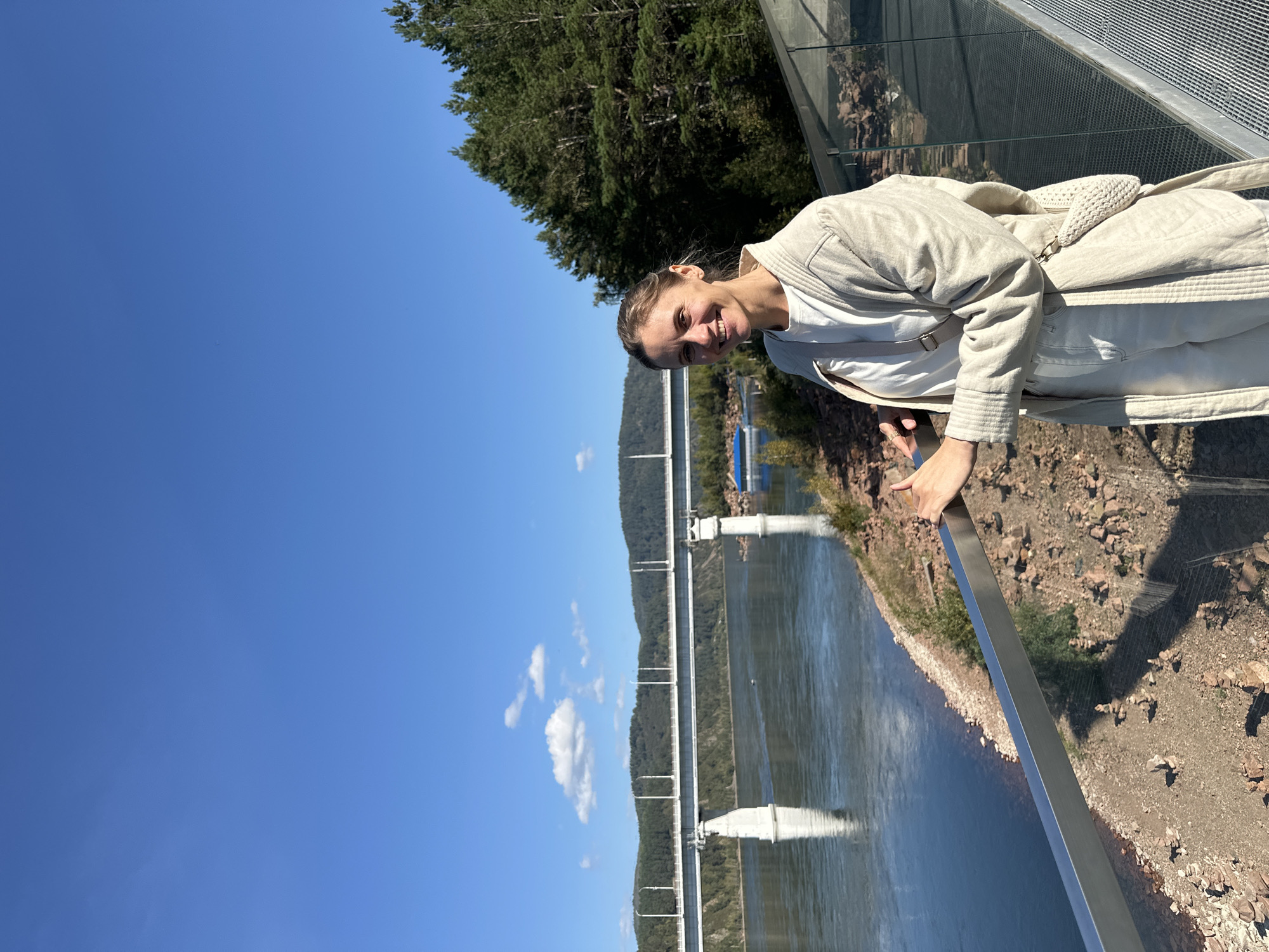 A woman with blonde hair pulled back smiles at the camera. She is wearing a white t-shirt, white pants, and a light-colored, textured jacket. A crossbody bag with a woven pouch is slung over her shoulder. She stands on a modern viewing platform with a metal railing and glass barrier. In the background, a wide river flows, and a large white bridge spans across it. Hills covered in dark green trees rise on the opposite bank. The sky is a clear, bright blue with a few wispy white clouds. The ground on the viewing platform and along the riverbank is covered with small rocks and pebbles, with some cairns formed from stacked stones visible near the water.