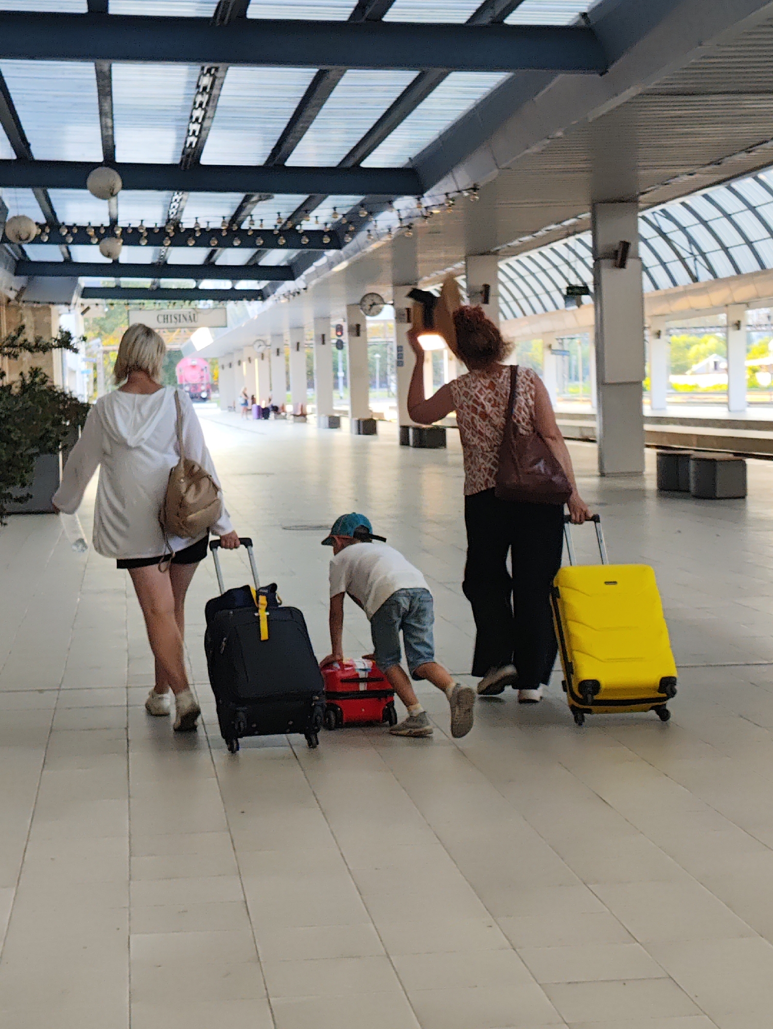 The image shows a view from the covered platform of the Chișinău railway station. The covered walkway has a modern design with a translucent roof supported by dark metal beams. Lights are strung along the underside of the roof. In the background, a red train is visible on the tracks.
In the foreground, three people are walking away from the camera. On the left is a woman with blonde hair, wearing a white hooded jacket and dark shorts. She is carrying a bottle of water in her left hand and pulling a black rolling suitcase with her right hand. A tan backpack is slung over her right shoulder. To her right is a young boy wearing a white t-shirt, blue shorts, and a blue cap. He is leaning down, pulling a small red rolling suitcase. To the right of the boy is a woman with curly brown hair, wearing a patterned sleeveless top and dark pants. She is holding the handle of a bright yellow rolling suitcase with her right hand and has a dark brown bag slung over her left shoulder. Her left hand is raised, holding a hat. The floor is paved with light-colored rectangular tiles. In the middle distance, other passengers can be seen sitting on benches or walking.