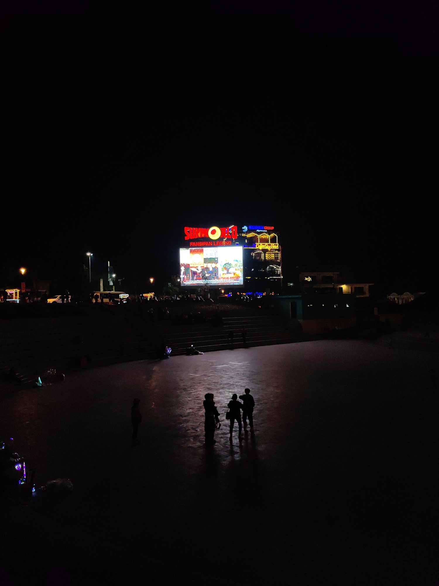 The image depicts a nighttime scene at a public plaza within the Sun World Fansipan Legend complex in Sa Pa, Vietnam. A large LED screen dominates the background, displaying colorful animated content and featuring prominent red and yellow signage for "SUNWORLD" and "FANSIPAN LEGEND." Below the screen, a crowd of indistinct figures is gathered on multiple tiers of steps and an elevated area, observing the display.
In the midground-left, a white car and a white bus or van are visible on a street, alongside streetlights. The foreground consists of a vast, dimly lit, paved surface. Five human figures are present in this foreground area, appearing as dark silhouettes against the faint reflections on the ground. Four figures are grouped closely together in the center-right, while a single figure stands further left. Near the isolated figure on the left, several objects on the ground emit subtle, colorful lights, primarily blue and green. The overall environment is a bustling public space characterized by artificial illumination against a dark sky.