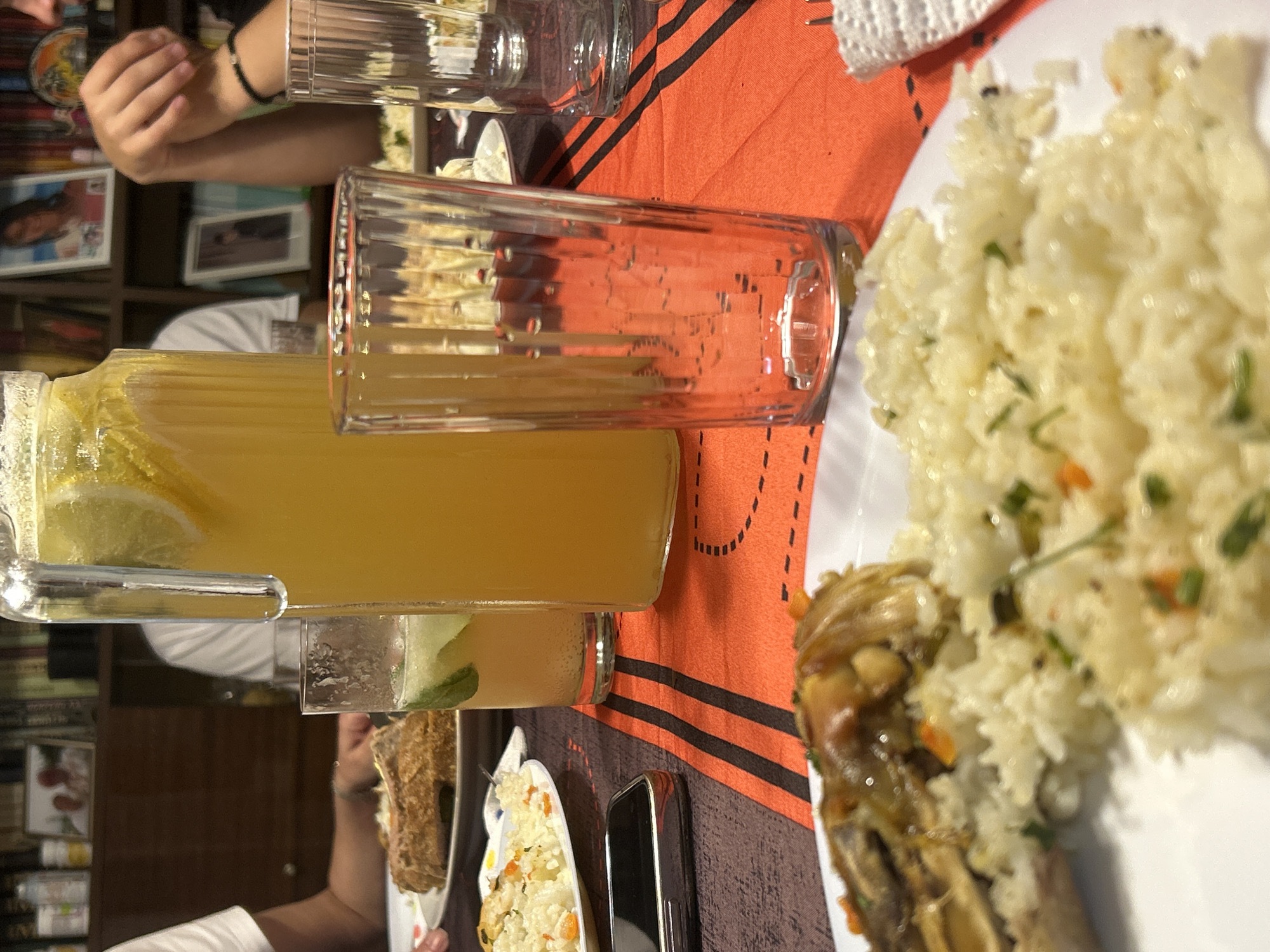 The image depicts an indoor dining scene in Buhovo, Bulgaria. A rectangular table, covered by a tablecloth featuring orange and dark brown stripes, is set for a meal.
In the foreground, a white plate on the lower left contains a portion of white rice garnished with green herbs and small orange pieces. Partially obscured on this plate is a large, irregularly shaped brown food item. To the right of this plate, a silver-edged smartphone is placed face down on the tablecloth. A tall, empty, clear glass with vertical fluting stands in the center foreground.
Dominating the center of the table is a large, clear glass pitcher filled with a yellow liquid, likely lemonade, containing several lemon slices and ice. To the left of the pitcher, a clear glass holds a lighter yellow-green beverage with visible slices of lime or cucumber and mint leaves, exhibiting carbonation bubbles. Further left, a white plate holds a large, crusty, brownish food item, possibly a loaf of bread or pastry.
On the right side of the table, another empty, fluted clear glass is present, and a white paper napkin or tissue is partially visible next to a metal fork. Another white plate with a portion of white food is partially visible beyond this glass.
Multiple individuals are present but largely out of frame. On the left, an arm wearing a white short-sleeved shirt is visible. Behind the pitcher, the torso and arm of an individual in a white short-sleeved shirt are discernible. On the right, an arm with a dark bracelet on the wrist is visible, resting near the edge of the table.
The background consists of dark wooden bookshelves filled with books and various objects. Several framed photographs are displayed on the shelves, including a larger framed image of a person on the top right shelf, and smaller framed images on the upper left and middle right shelves.