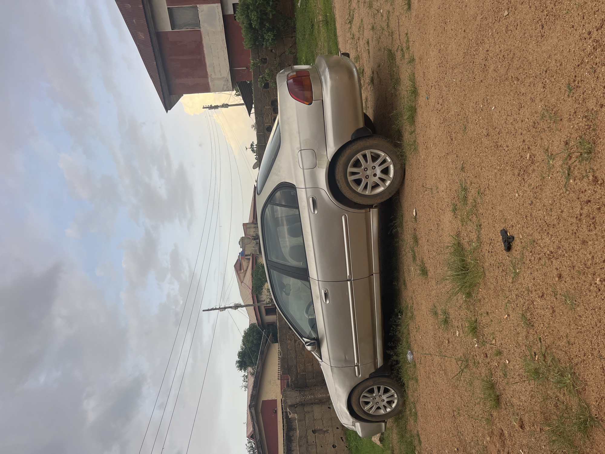 A silver four-door sedan is parked on a dirt road in front of a low brick wall and residential buildings in Jos, Nigeria. The sky is overcast with scattered clouds. The car is positioned parallel to the wall, facing right in the frame. In the foreground, the dirt road has patches of dry grass. A small, dark object, possibly a toy gun, lies on the ground near the car's rear tire. Behind the car, the brick wall is visible, with parts of buildings and a corrugated metal roofed structure further back. Telephone wires are strung across the sky.