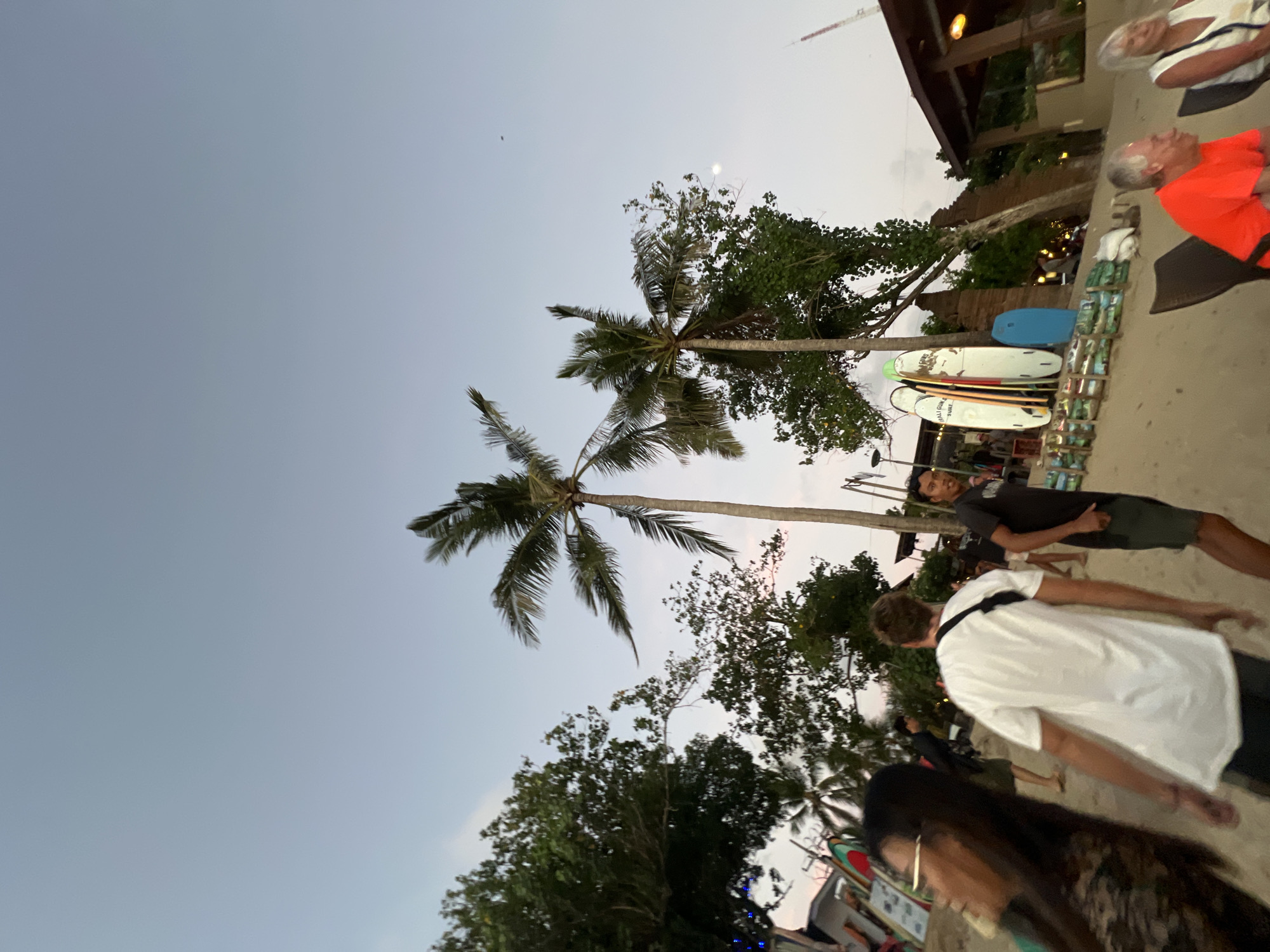 This image captures a scene on Kuta Beach in Kuta, Indonesia, during twilight. The sky is a pale blue with hints of pink and orange near the horizon. Several palm trees are scattered across the sandy beach, with their fronds silhouetted against the sky. In the foreground, a woman with long dark hair and glasses is partially visible, looking to her left. To her right, a man wearing a white t-shirt and dark shorts walks away from the camera. Further back, another man in a dark t-shirt and shorts stands facing the camera, with a group of surfboards propped up behind him. In the background, there are several beachside establishments with thatched roofs, illuminated by warm lighting. The overall atmosphere suggests a relaxed, late-day scene on a popular tourist beach.