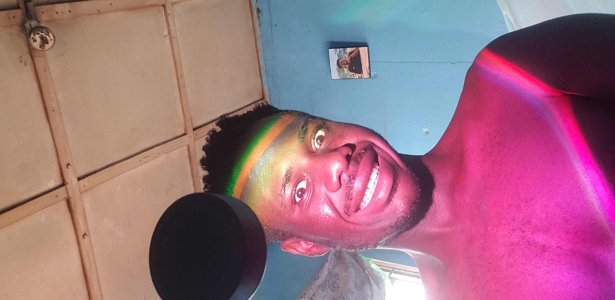 In Narayi, Nigeria, an individual is captured in a selfie-style photograph taken indoors. The person, a dark-skinned male with short, dark hair, is positioned centrally in the frame and is looking directly at the camera with a wide, smiling expression and wide eyes. A rainbow-colored light, possibly from a prism or light fixture, is cast across his forehead, cheek, and shoulder, also illuminating the wall behind him. He is wearing a pink-toned light that covers his upper body. To the left of his head, a black, circular object is partially visible. The background consists of a light blue wall and a ceiling with a grid pattern, featuring what appears to be a security camera. A framed picture is mounted on the wall behind the individual. The overall impression is that of a candid and colorful portrait, with the light effects creating a dynamic and engaging visual.